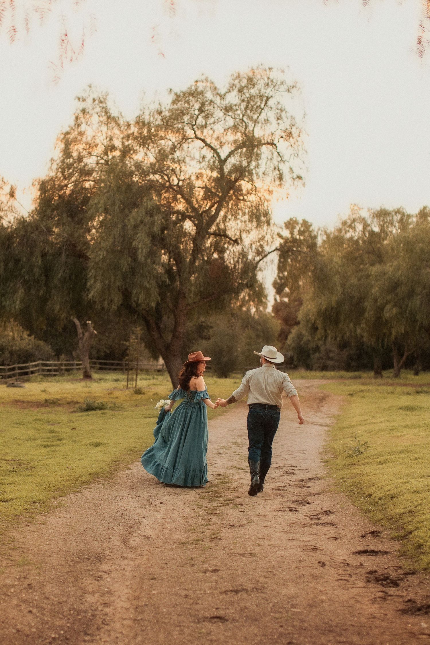 Couple walking hand in hand down a dirt path at sunset with a woman in teal dress and man in casual attire.