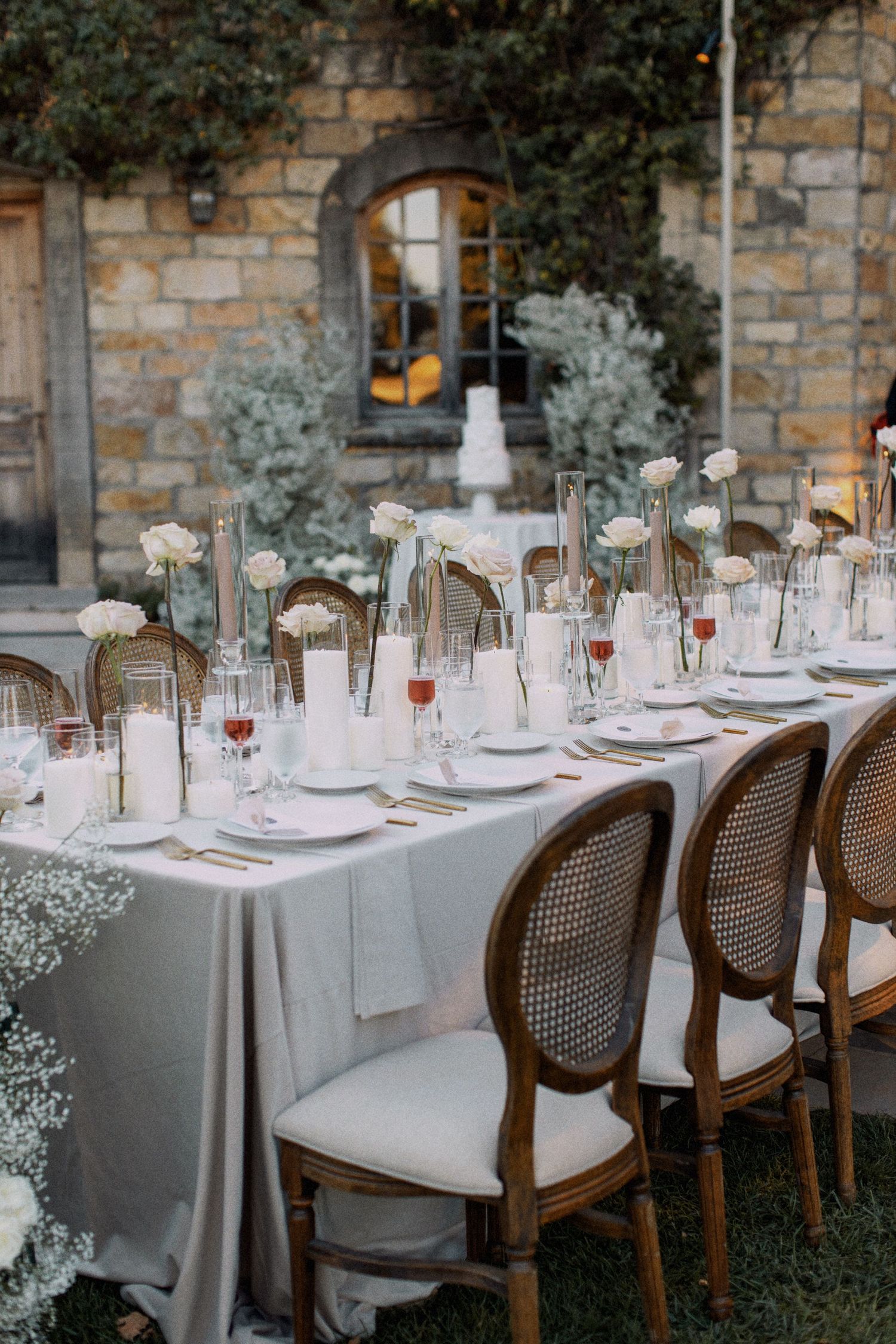 Elegant outdoor wedding reception table with white linens, floral centerpieces and cane-back chairs.
