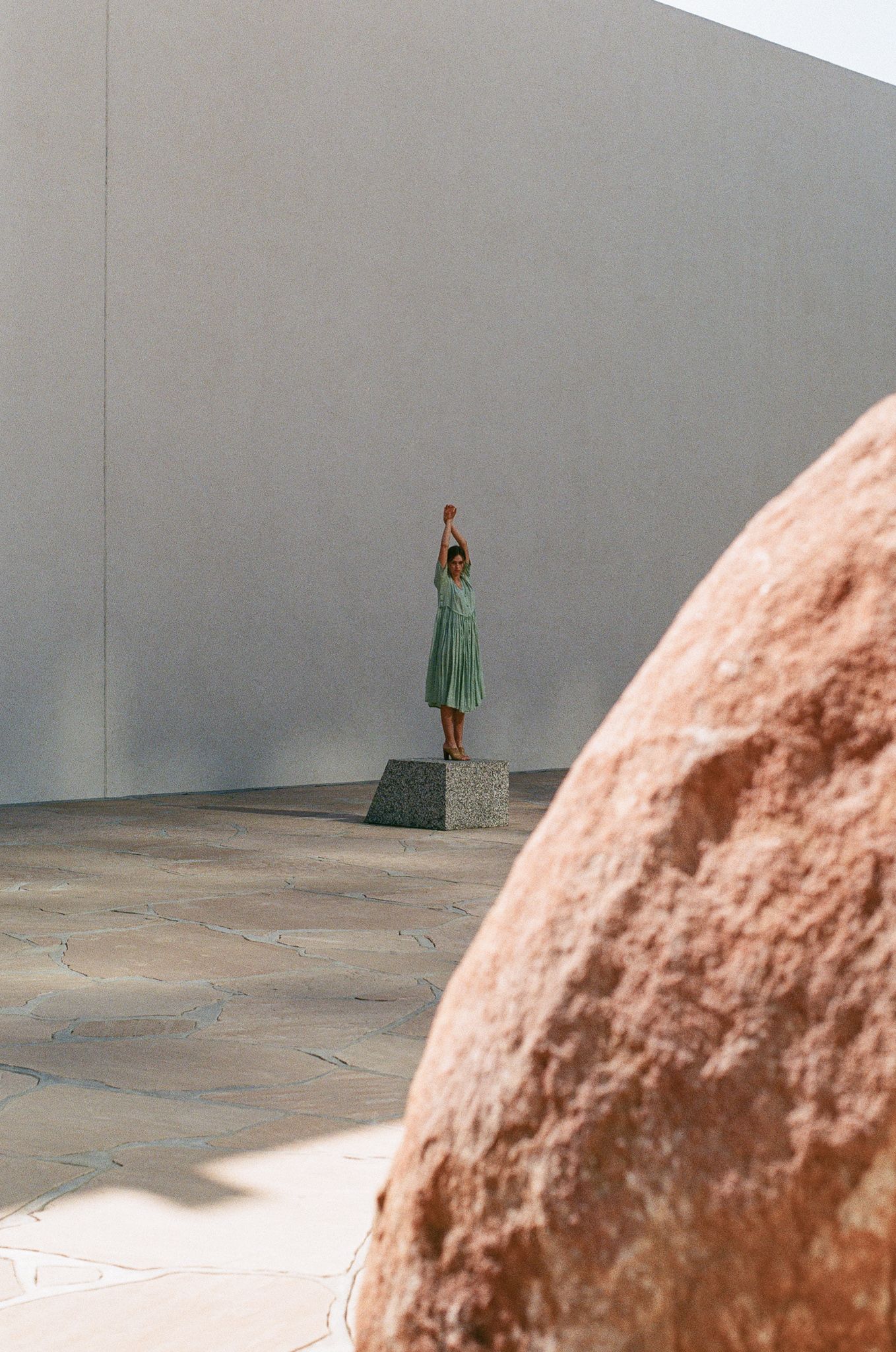 Person in green dress stands on pedestal near large stone wall in artistic composition.