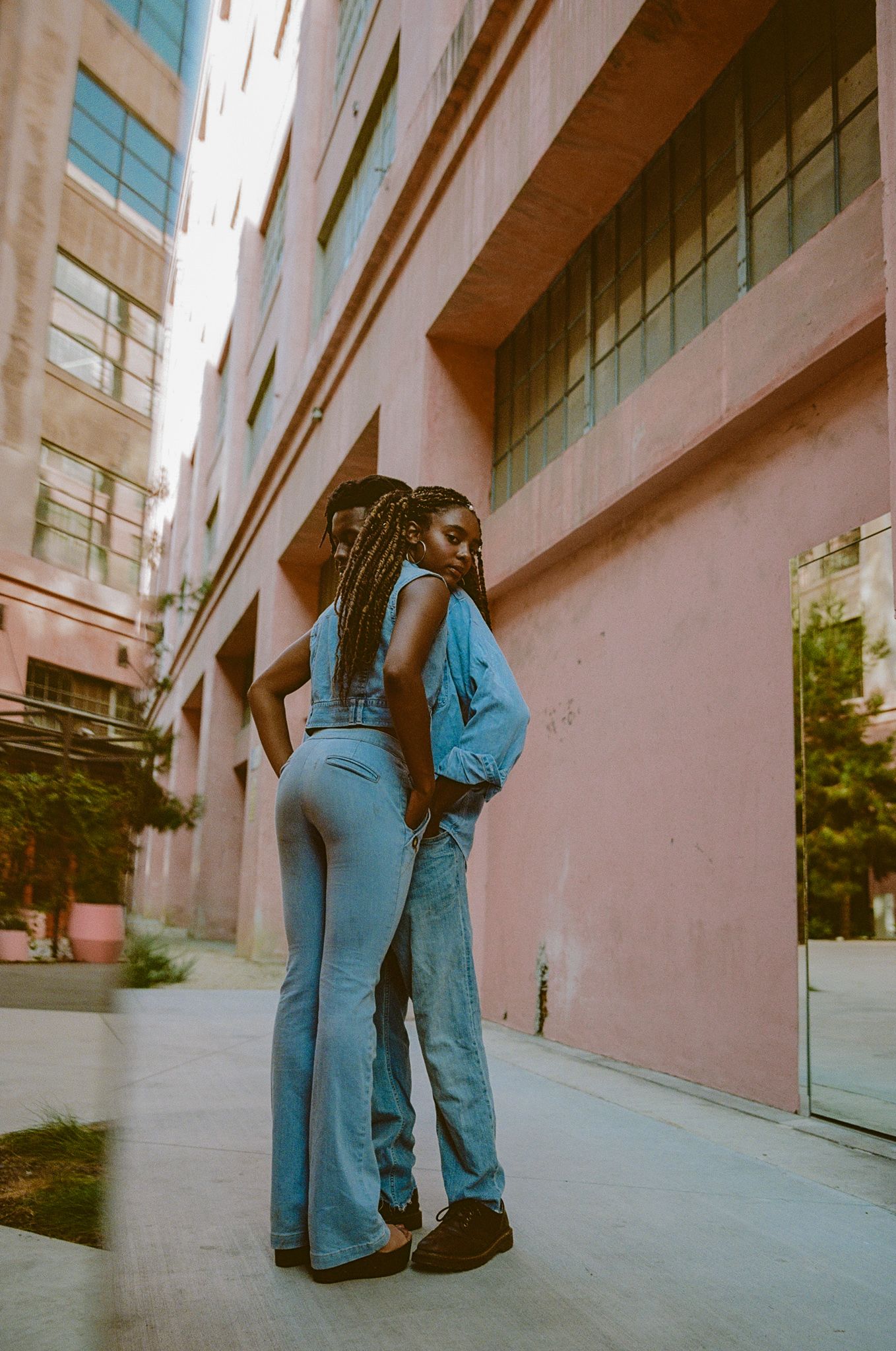A couple in matching denim outfits stands close together against a modern building exterior in an urban setting.