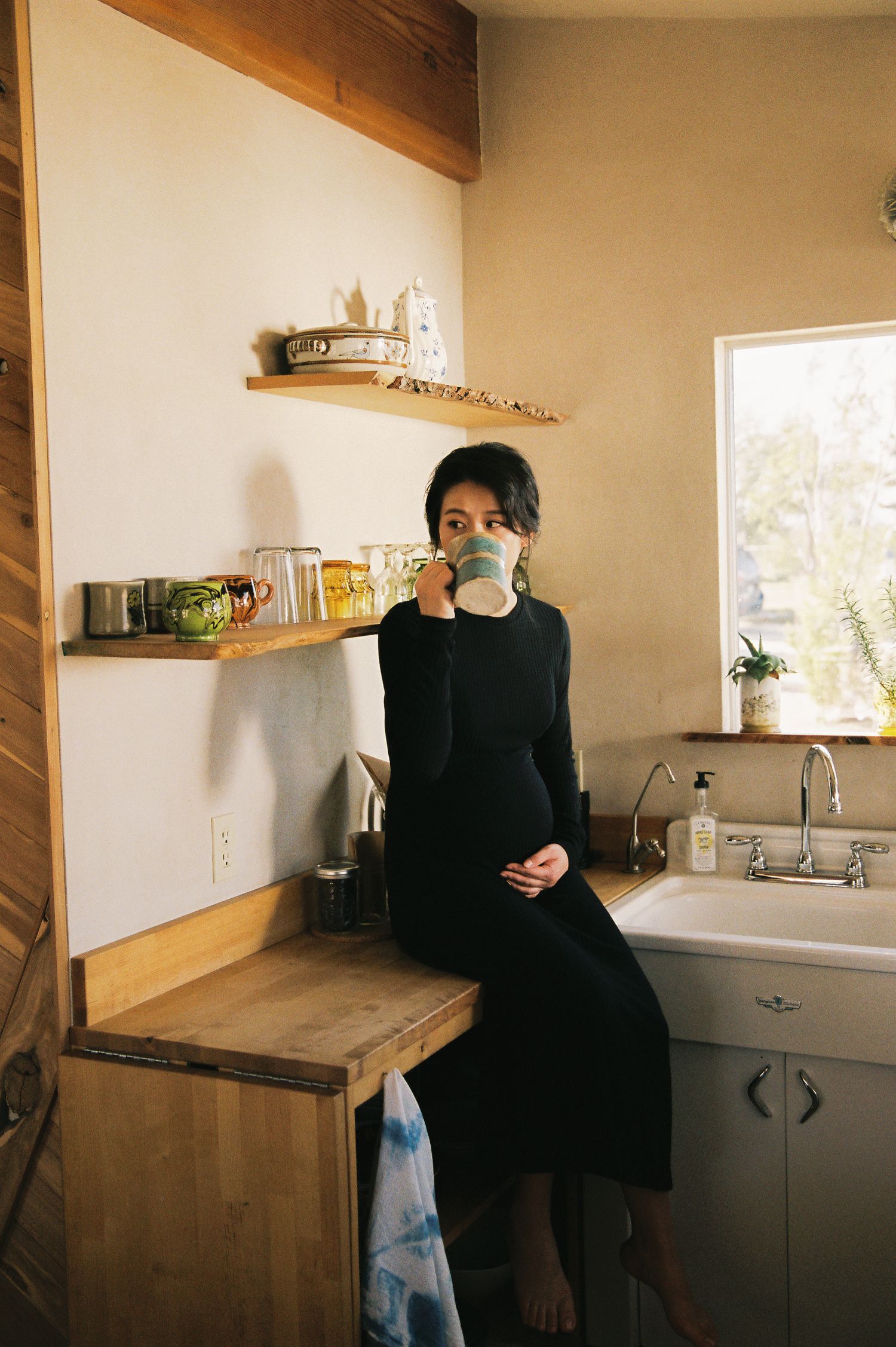 Person in black dress sipping coffee while sitting on wooden counter in rustic kitchen.