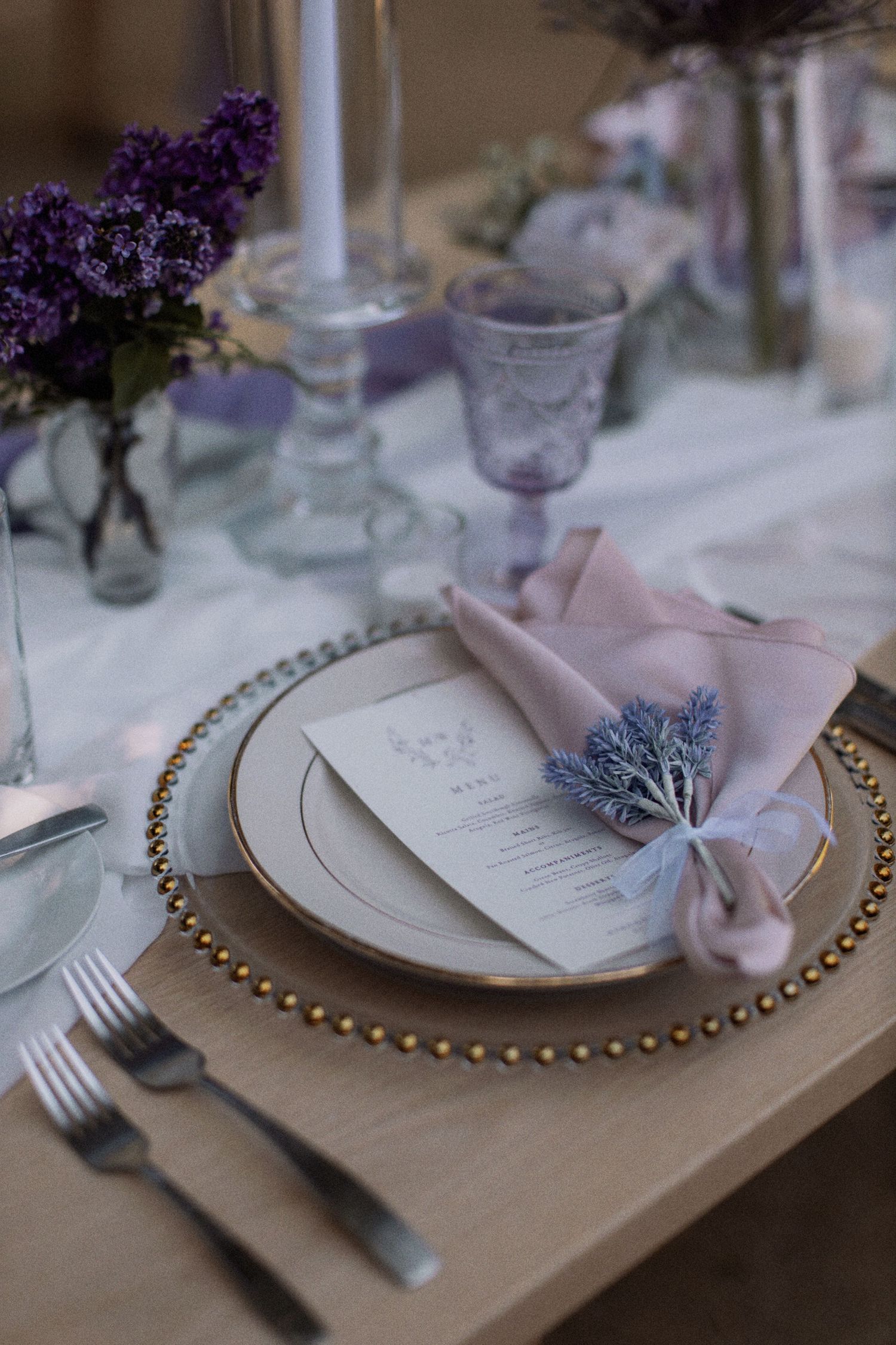 Elegant place setting with gold-rimmed charger plate, purple glassware, and lavender napkin decoration.