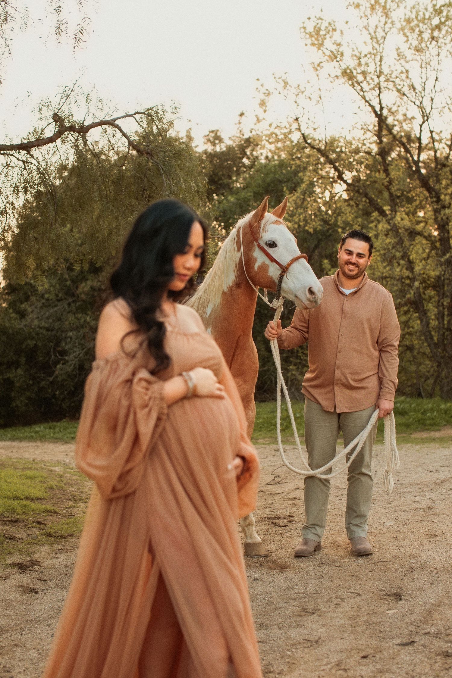A couple poses in a rustic outdoor setting at sunset with their white horse, wearing matching peach-colored outfits.