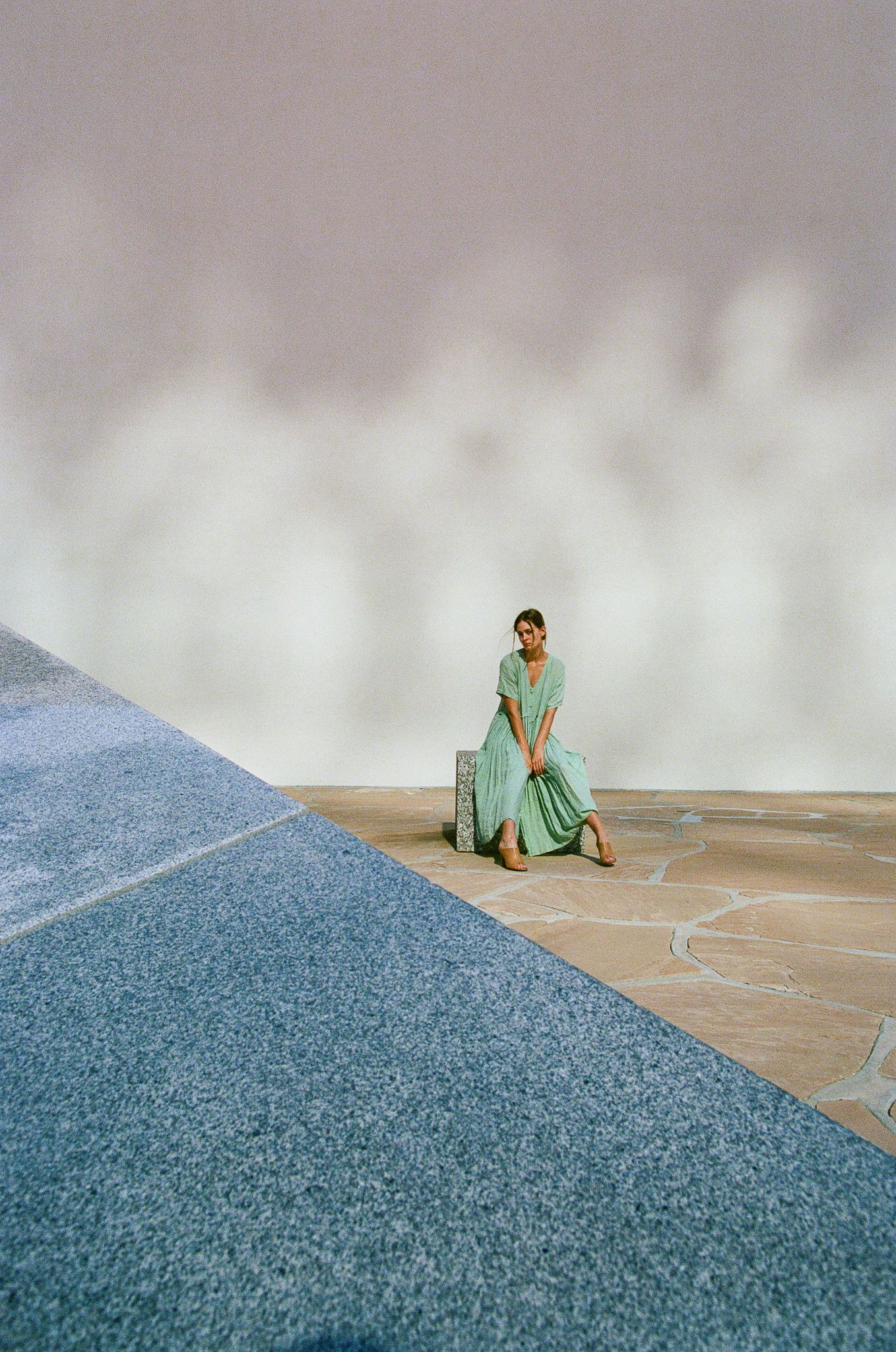A figure in a flowing mint green dress poses against a geometric backdrop with blue concrete and white walls.