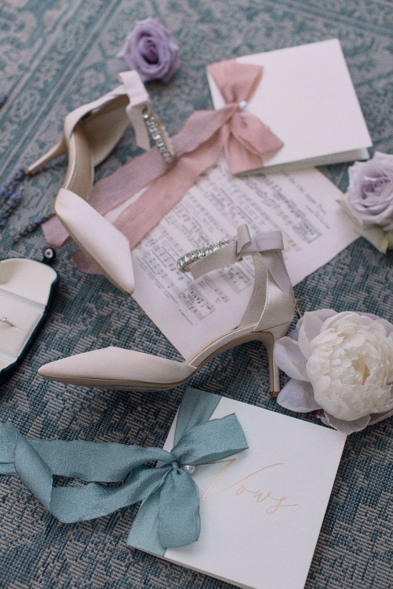 Elegant wedding accessories including white heels, roses, and ribboned invitations on a patterned surface.