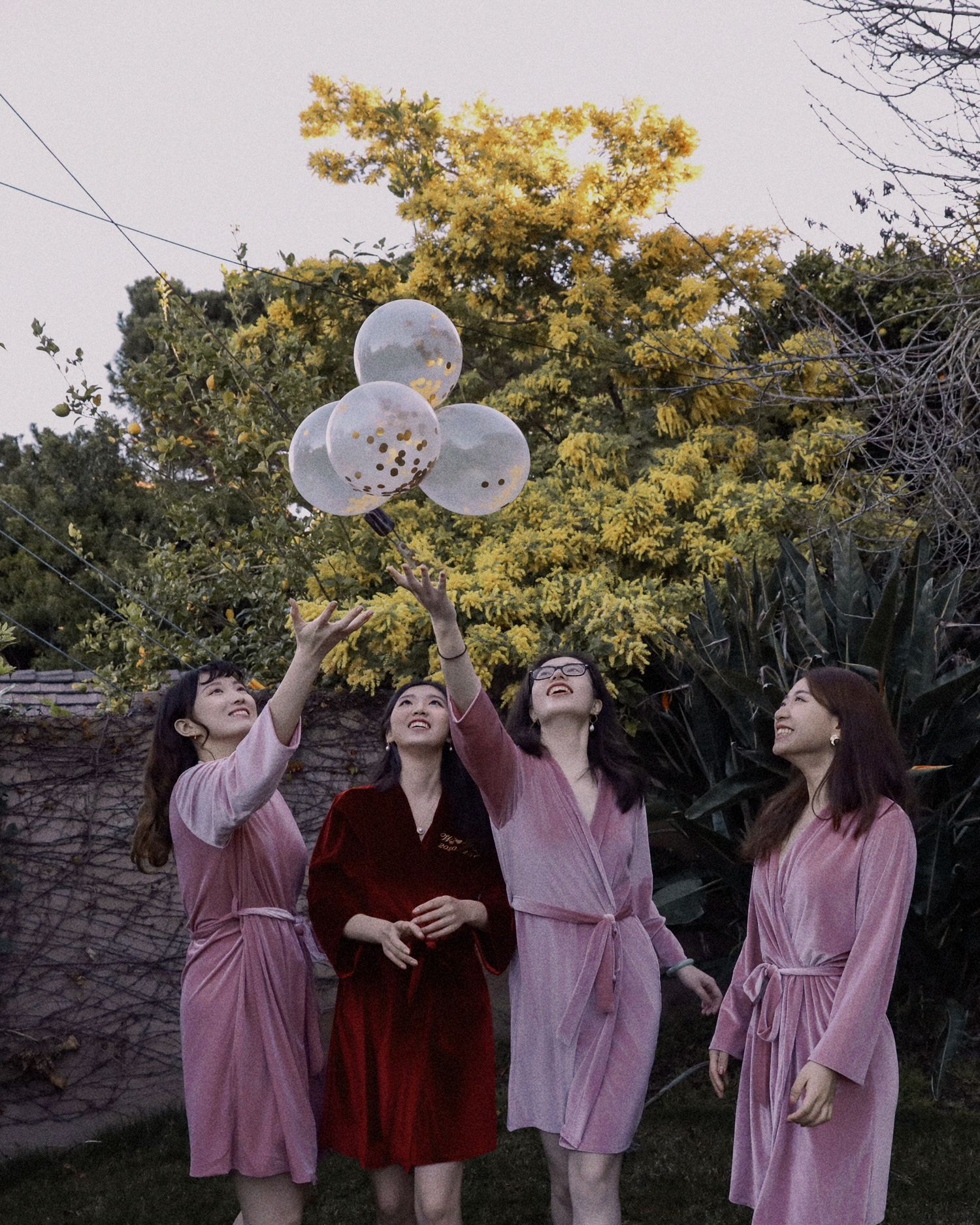 Bridesmaids in pink robes look up at floating balloons in garden setting.