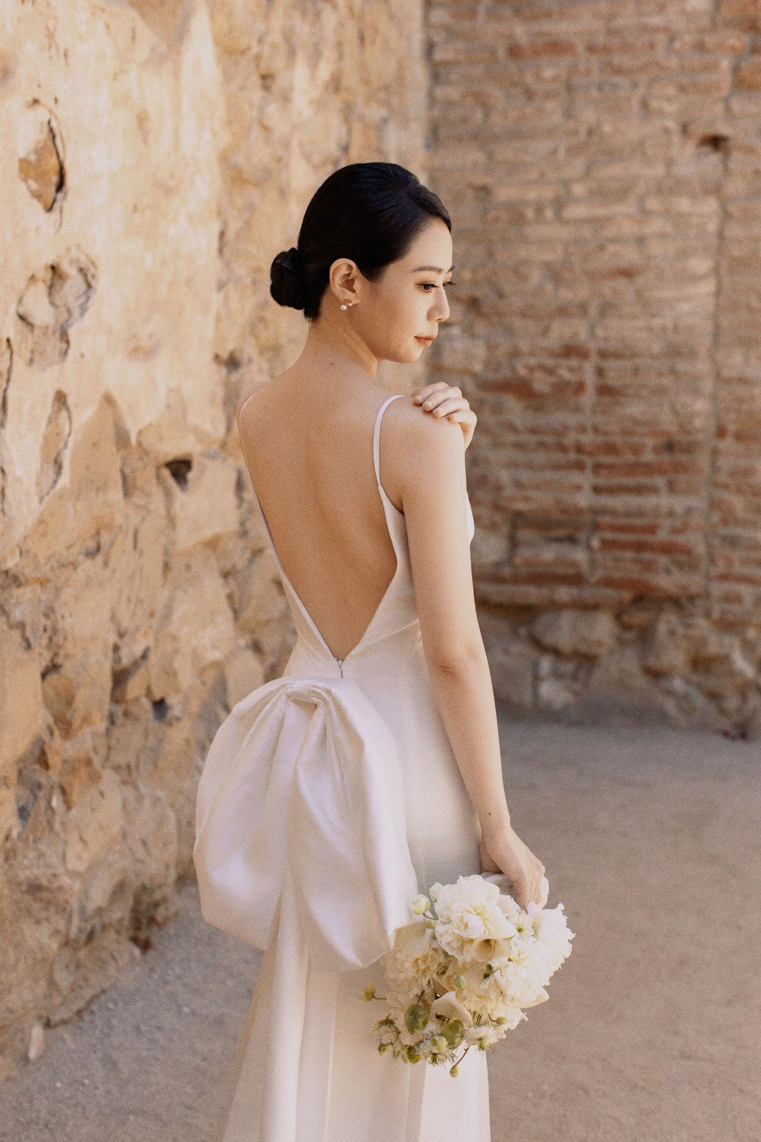 Elegant bride in backless white dress holds a bouquet against brick wall backdrop.