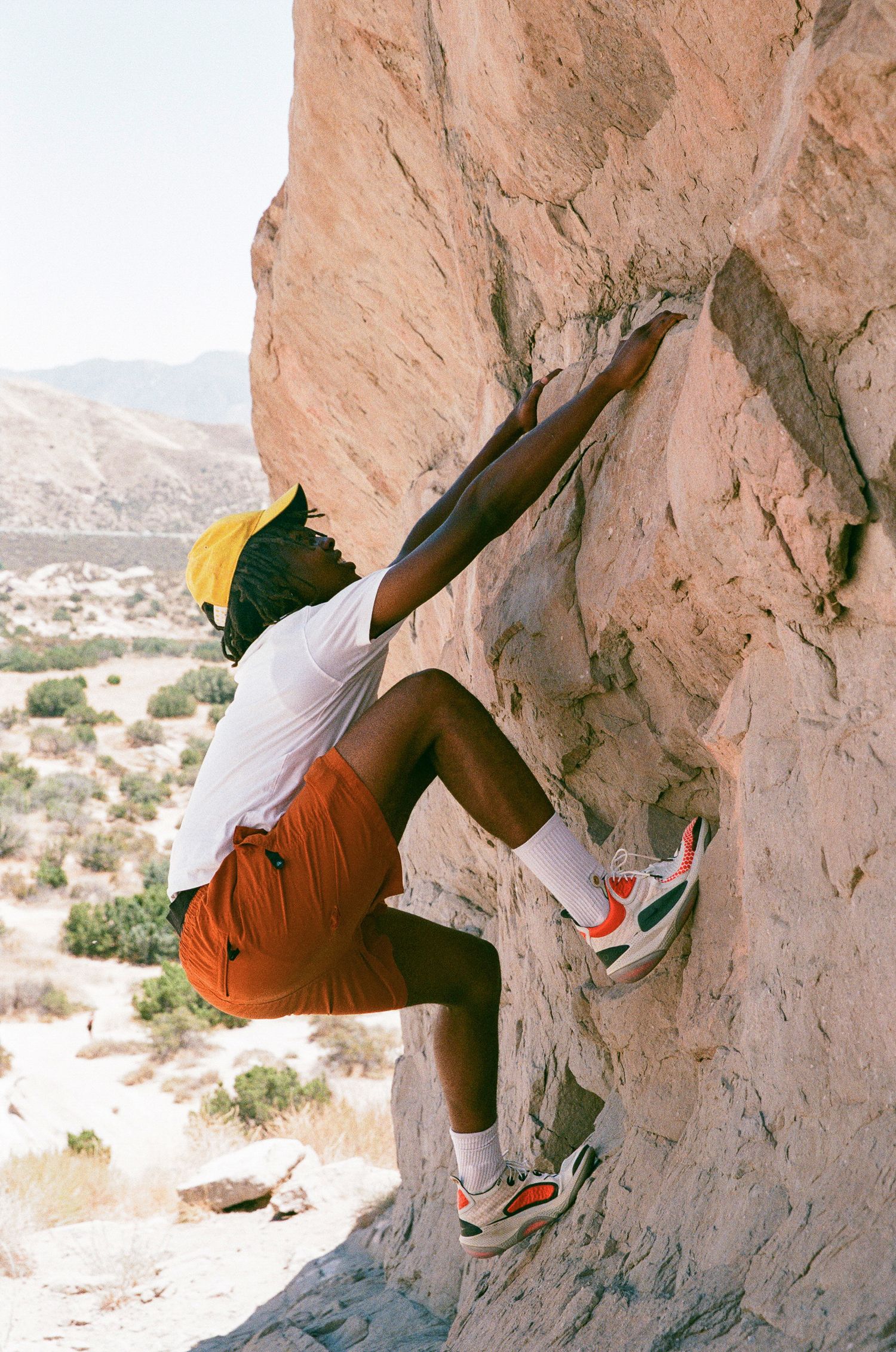 A rock climber in orange shorts tackles a challenging desert cliff face.