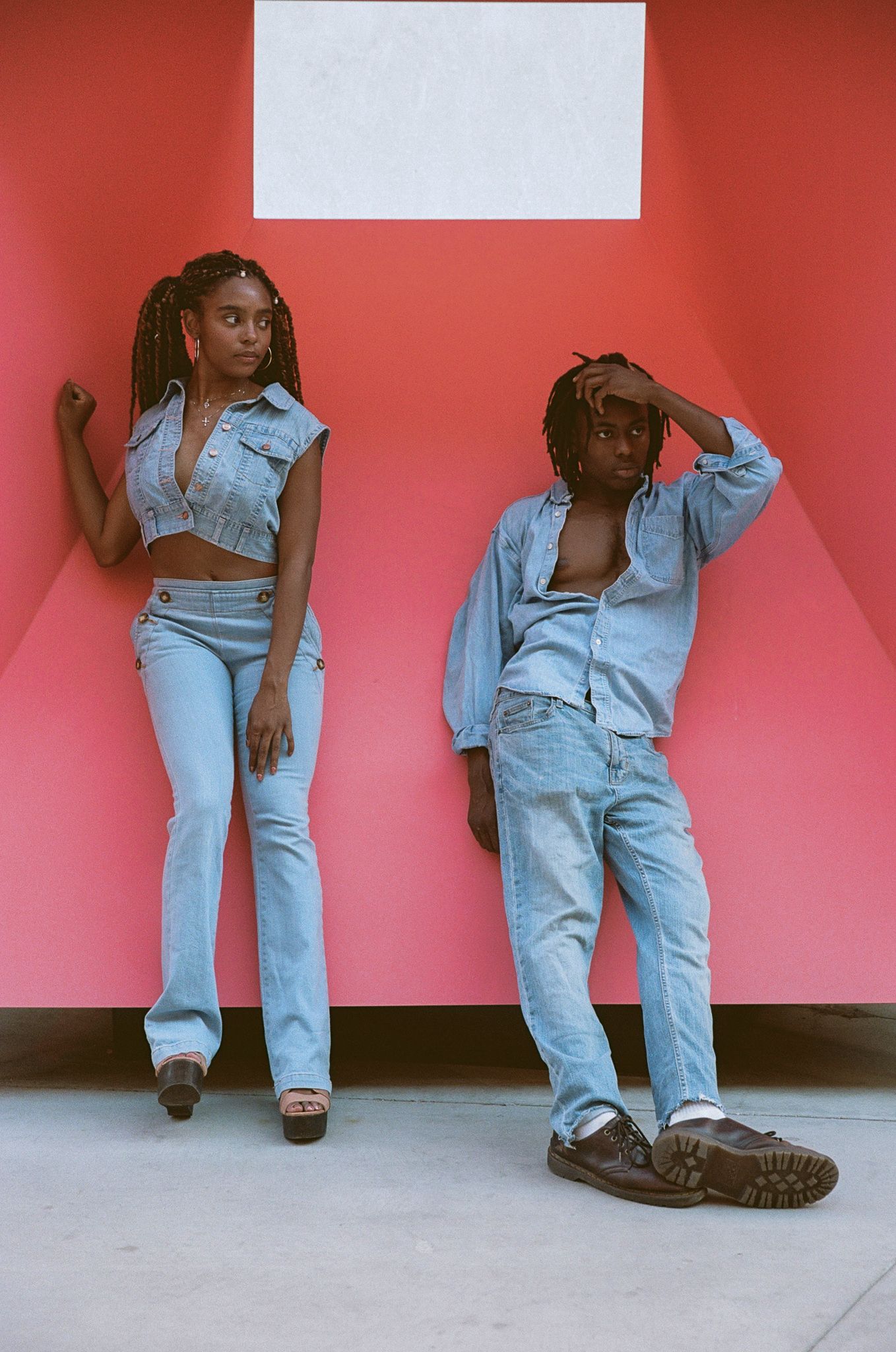 Two people in denim outfits pose against a coral pink wall, wearing casual blue jeans and denim shirts.