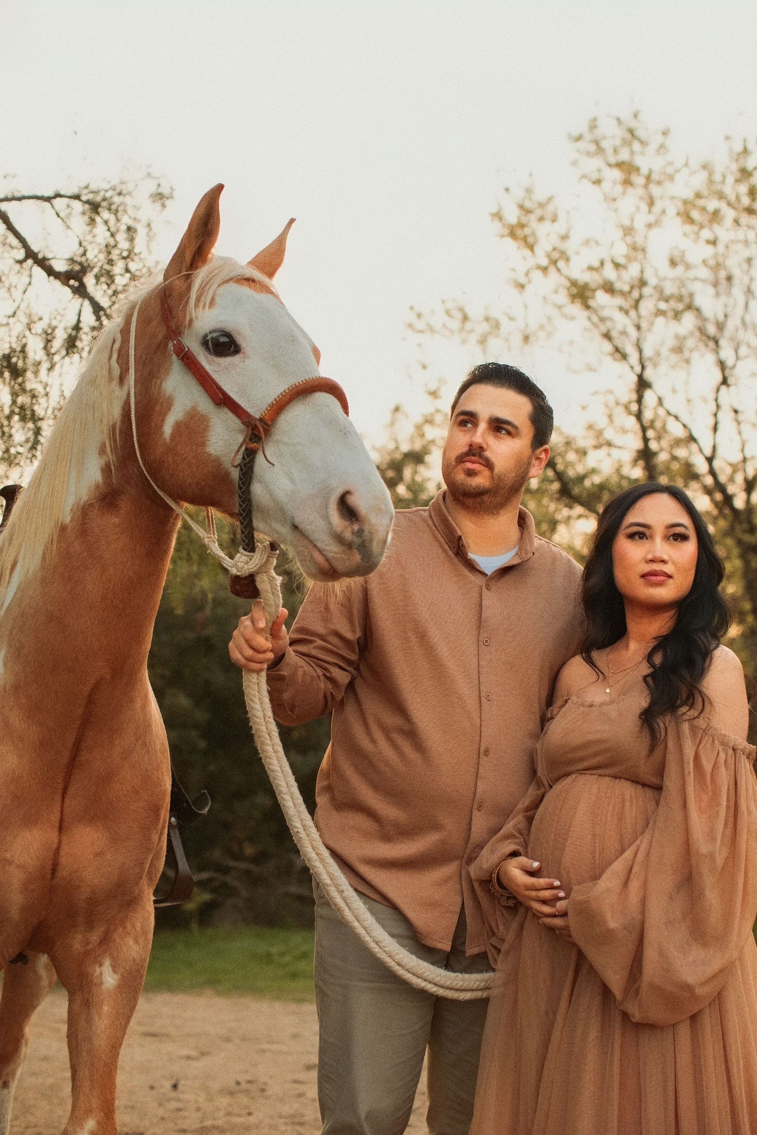 A couple in muted brown tones stands next to a light-colored horse in an outdoor autumn setting.