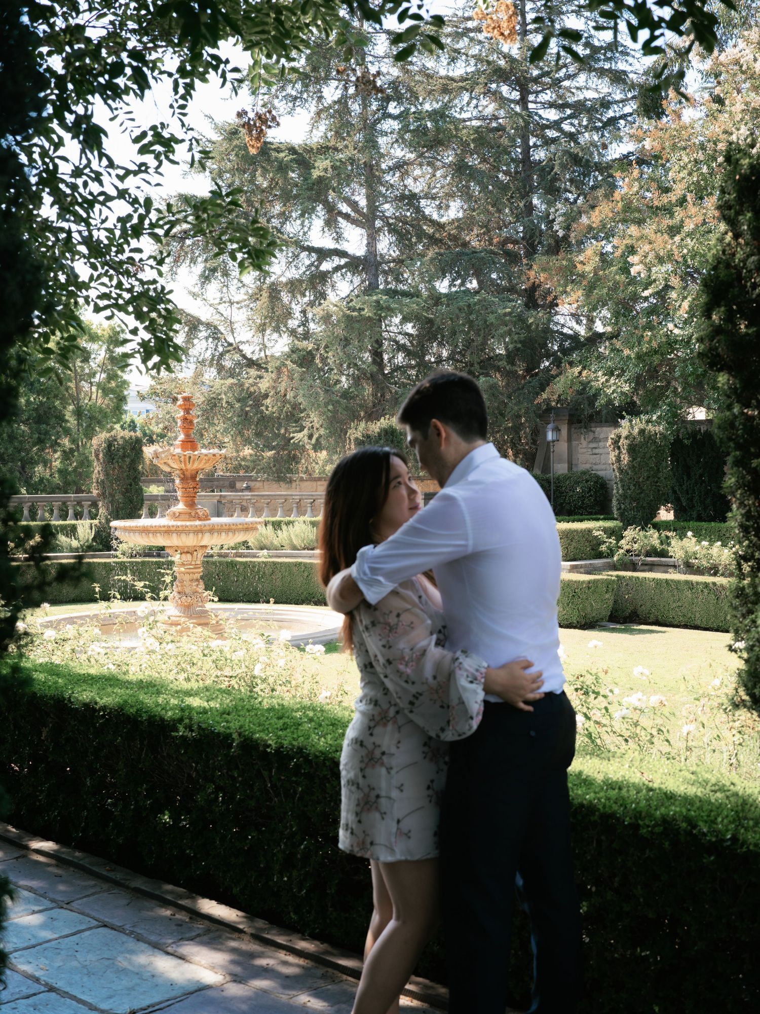 A couple embraces in a sunlit garden with a classical fountain in the background.