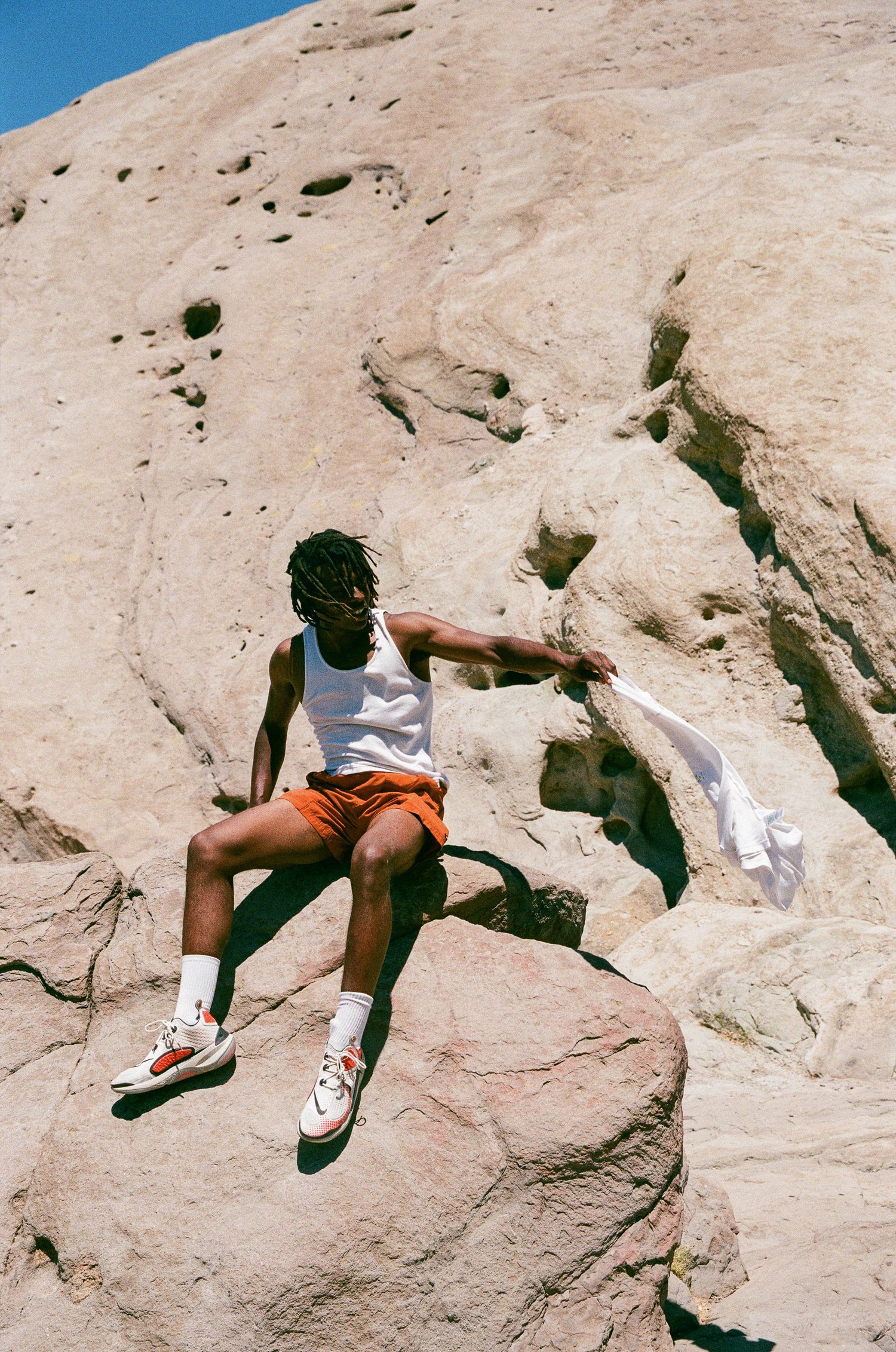 A person in a white tank top and orange shorts sits on a rocky cliff face.