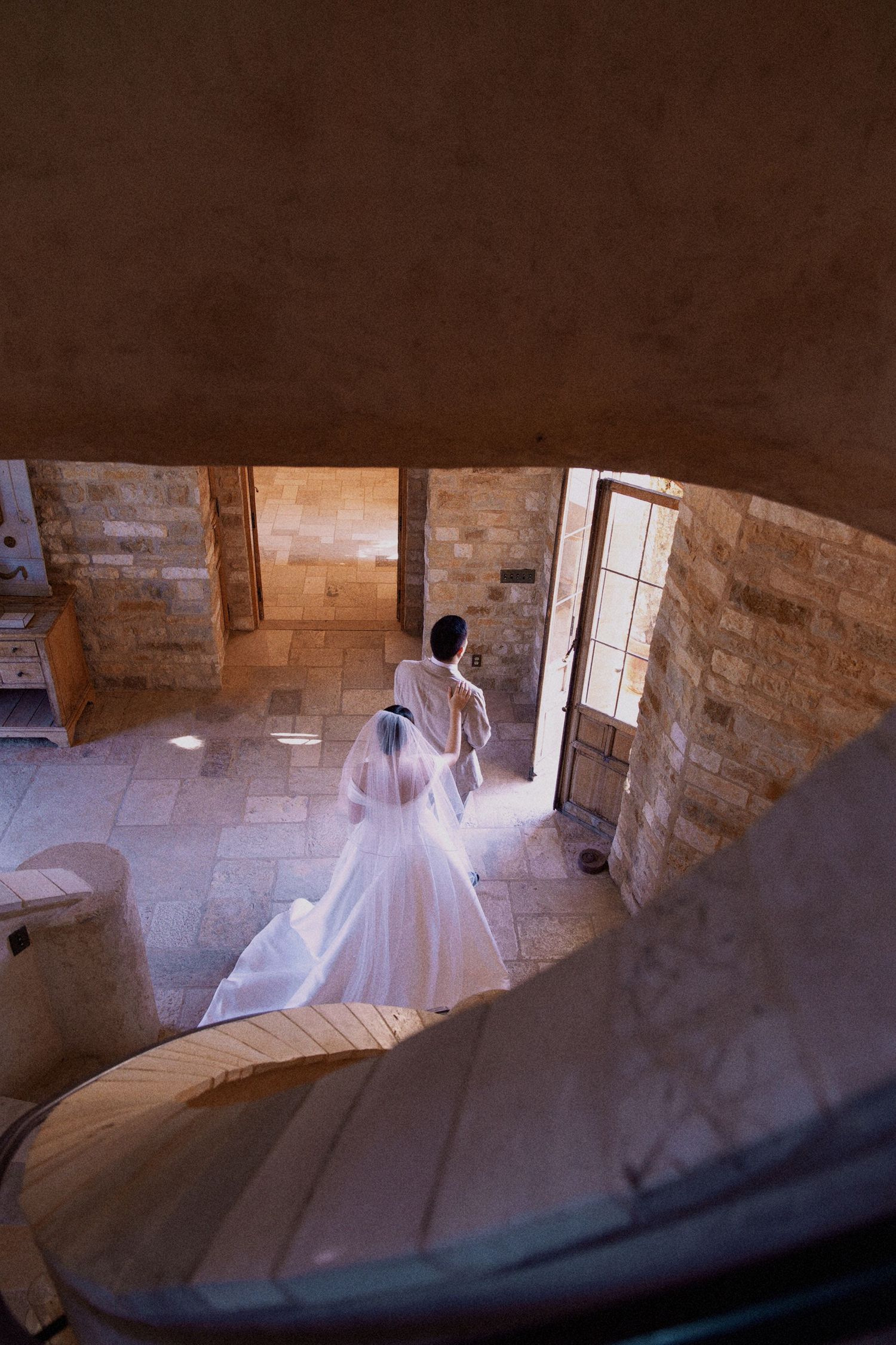 A bride in a white wedding gown descends a stone spiral staircase in a historic castle setting.