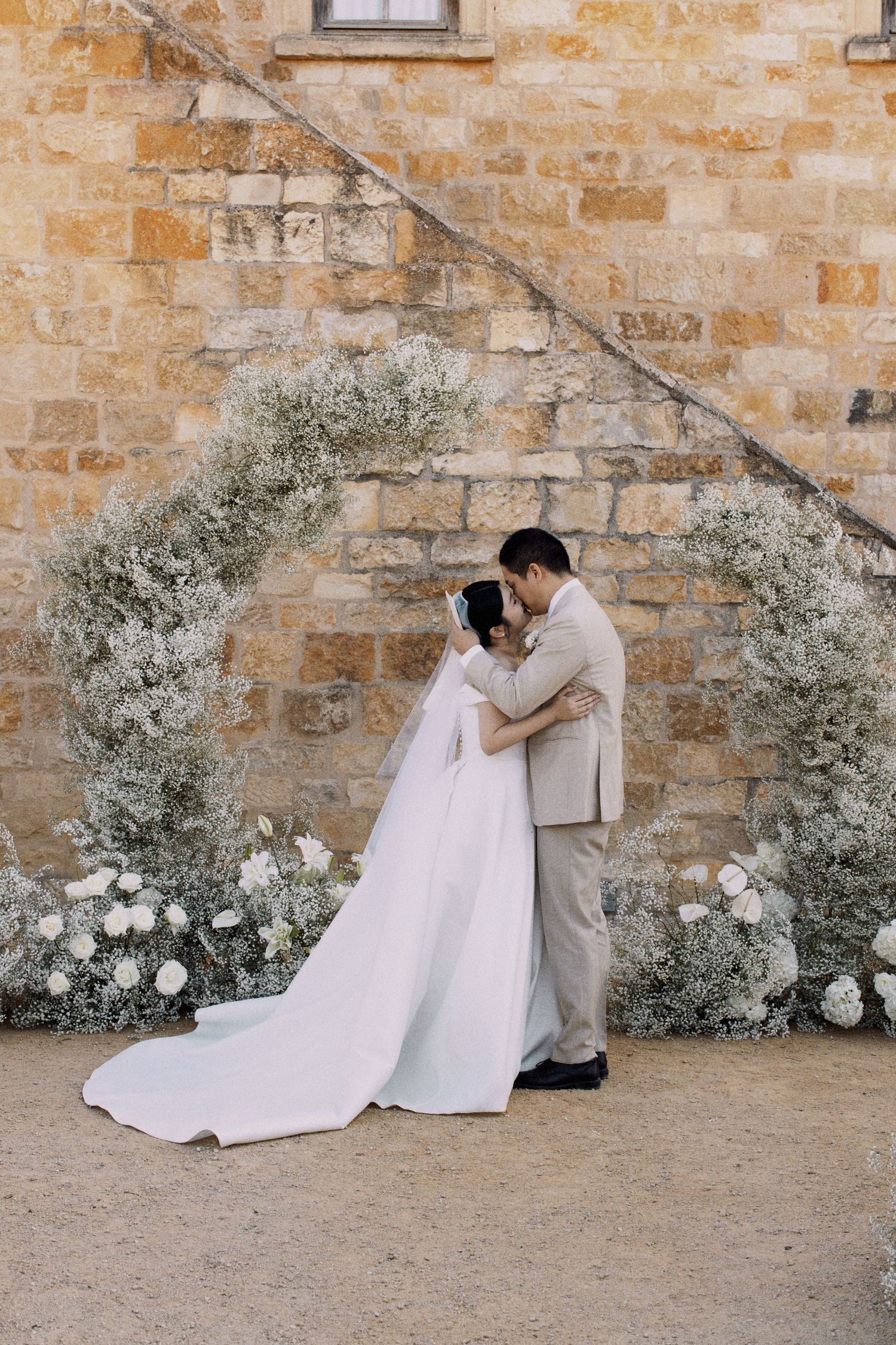 Elegant wedding couple embraces under a dreamy floral arch against rustic stone wall at golden hour.