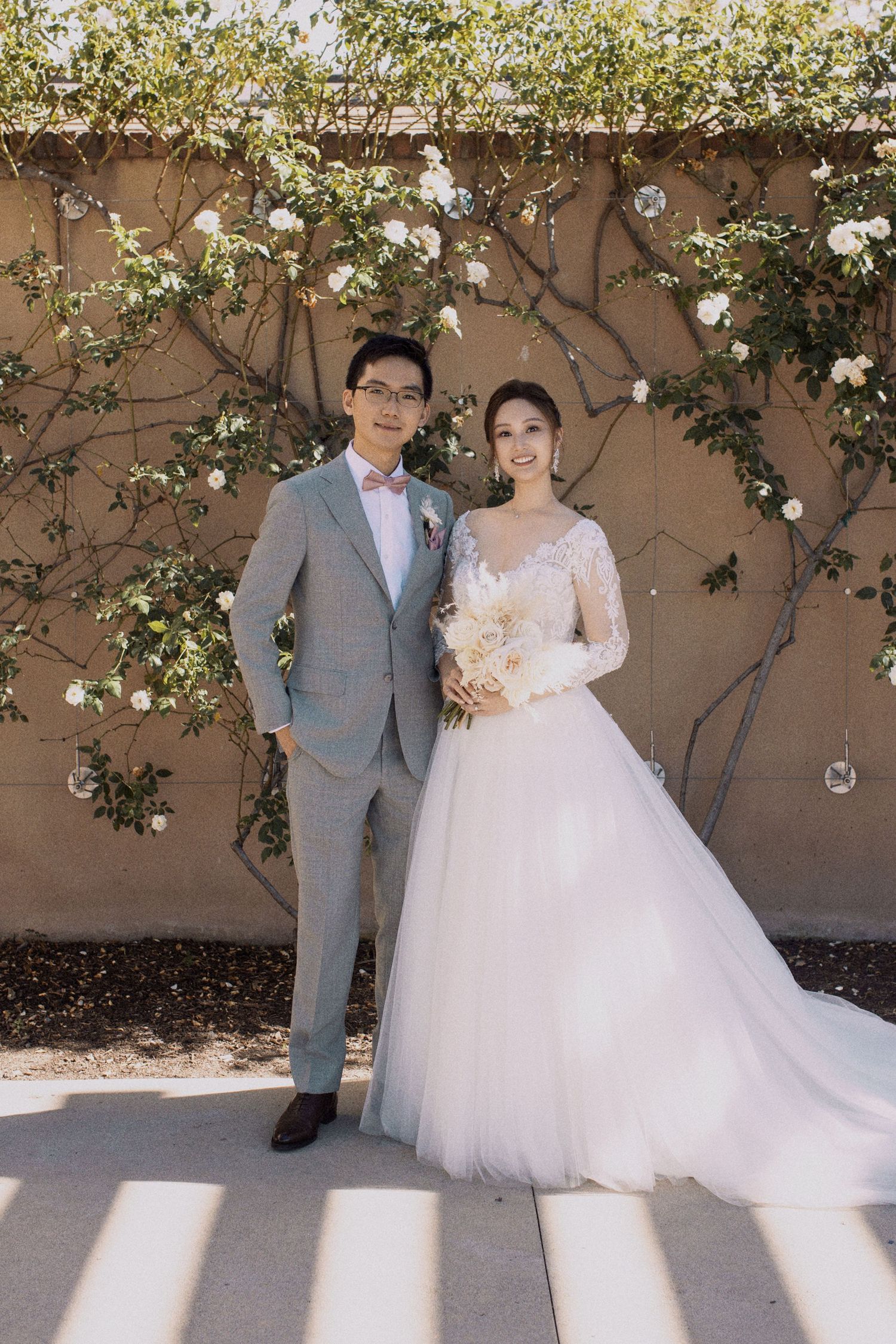 Elegant bride in lace gown and groom in grey suit pose against floral wall with white roses.