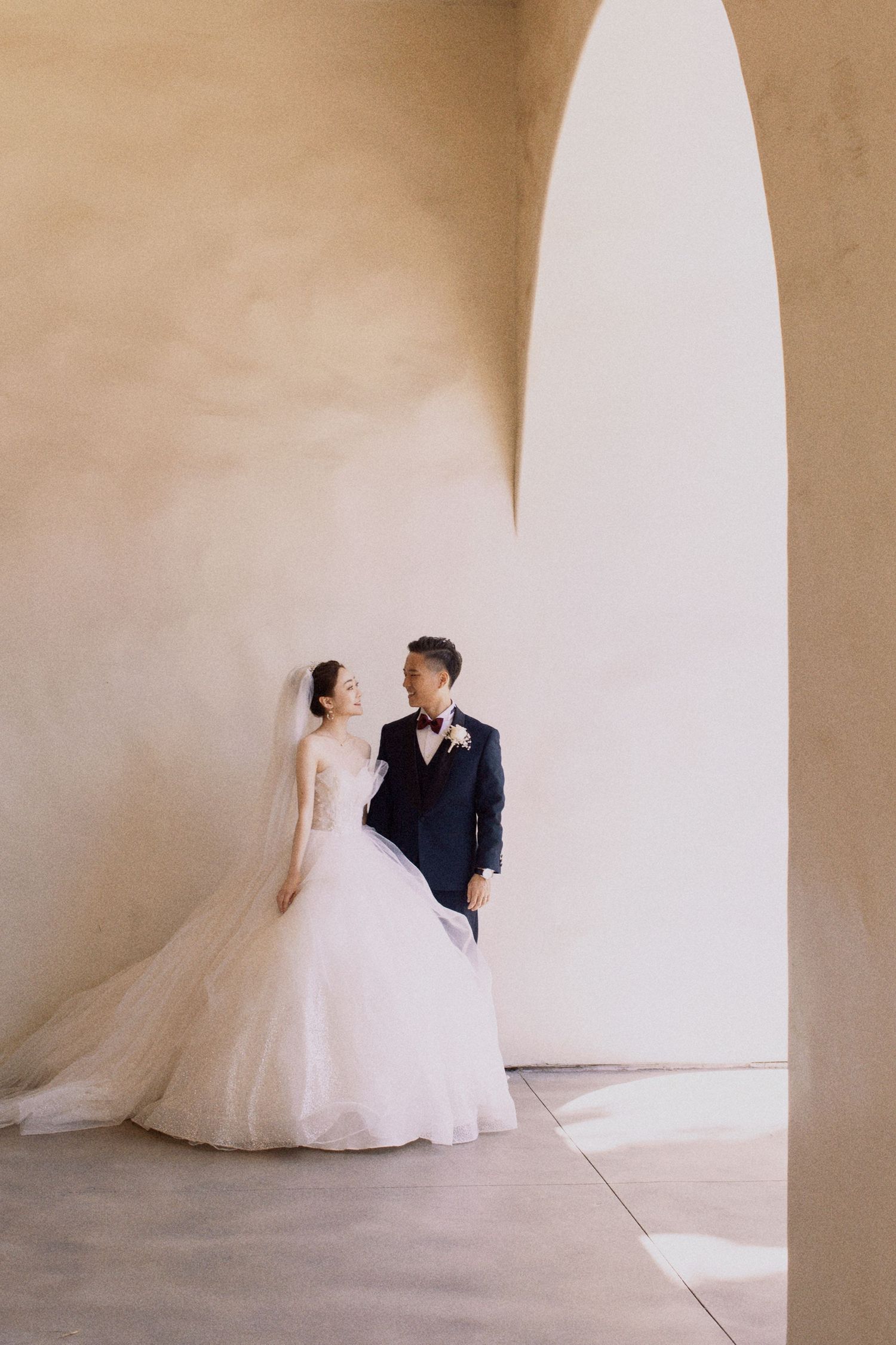 A wedding couple poses under an elegant archway in soft natural lighting.