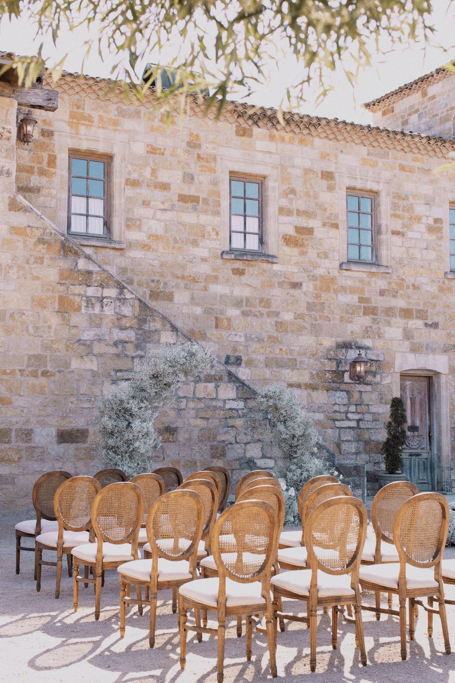 Elegant wooden chairs arranged for an outdoor ceremony against a rustic stone building facade.