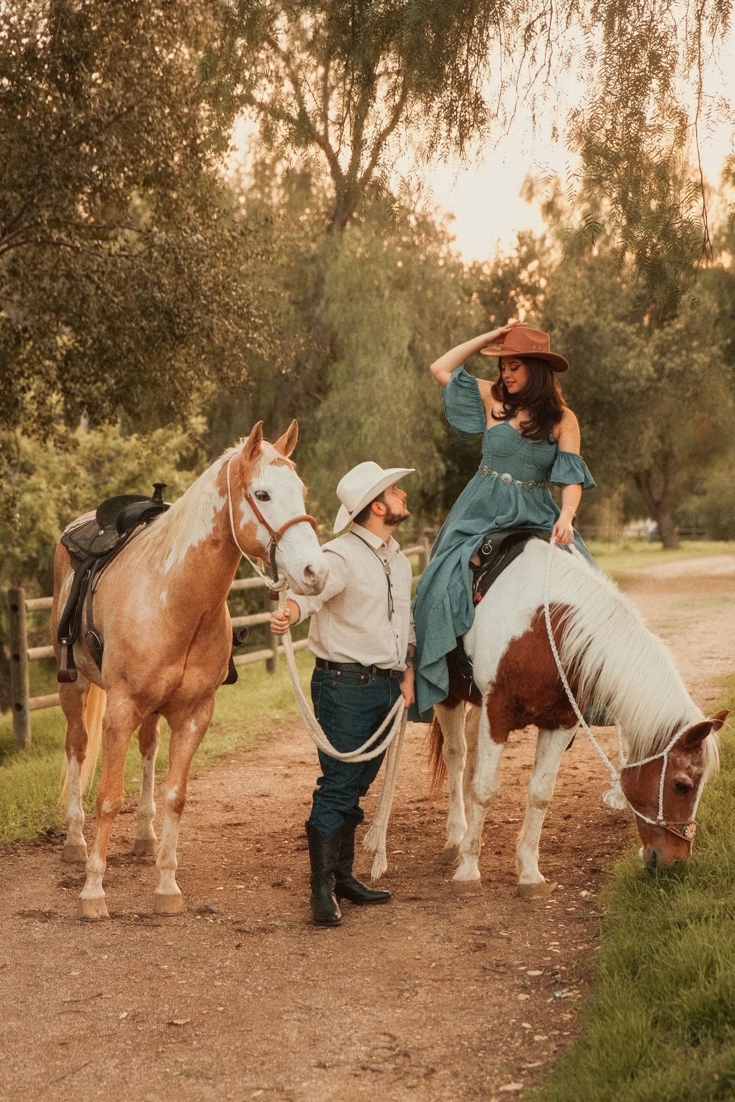 Two riders in casual western attire with hats enjoy a sunset trail ride on paint and palomino horses along a dirt path.