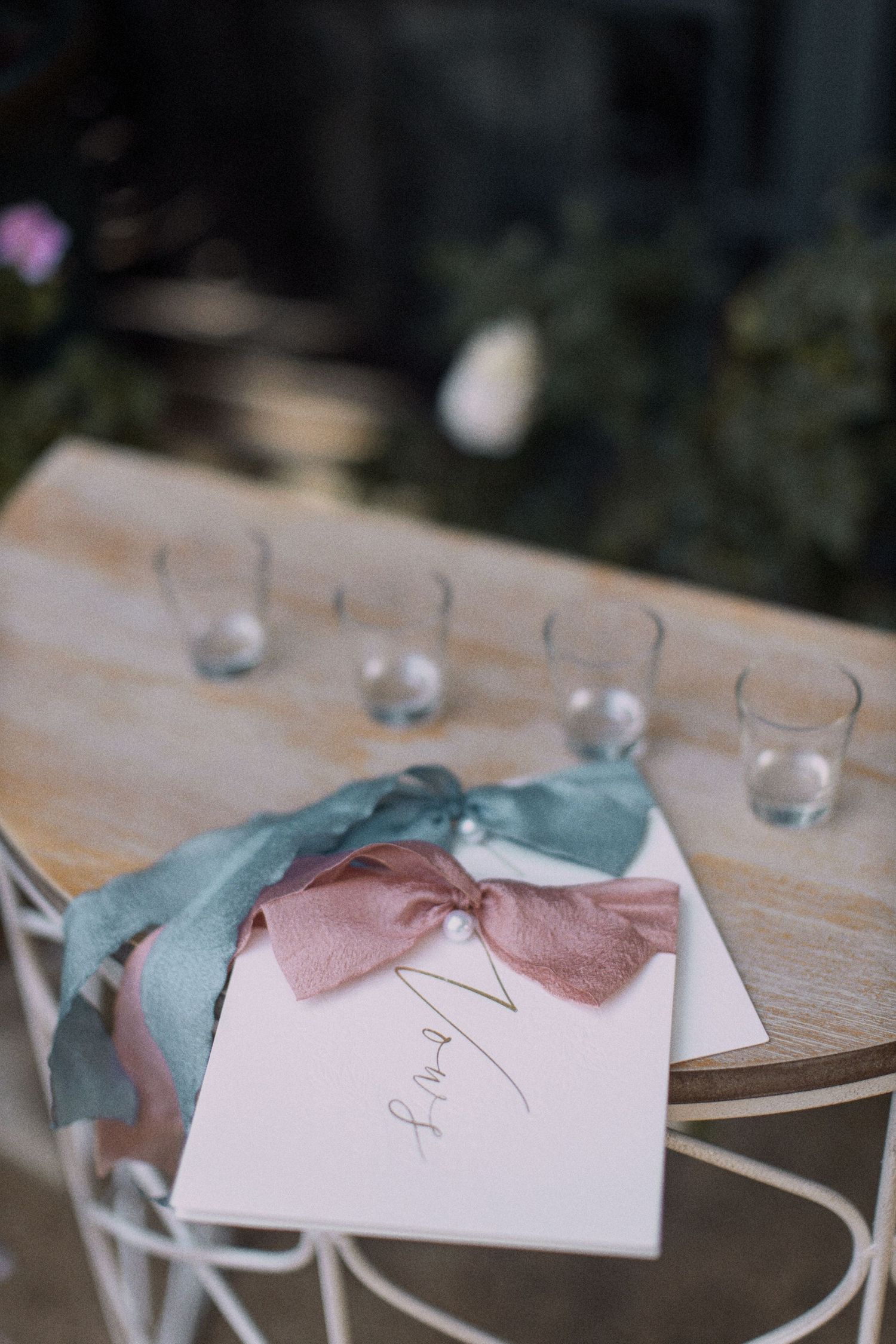 Wedding stationery with pink and sage ribbons on wooden table with glassware.