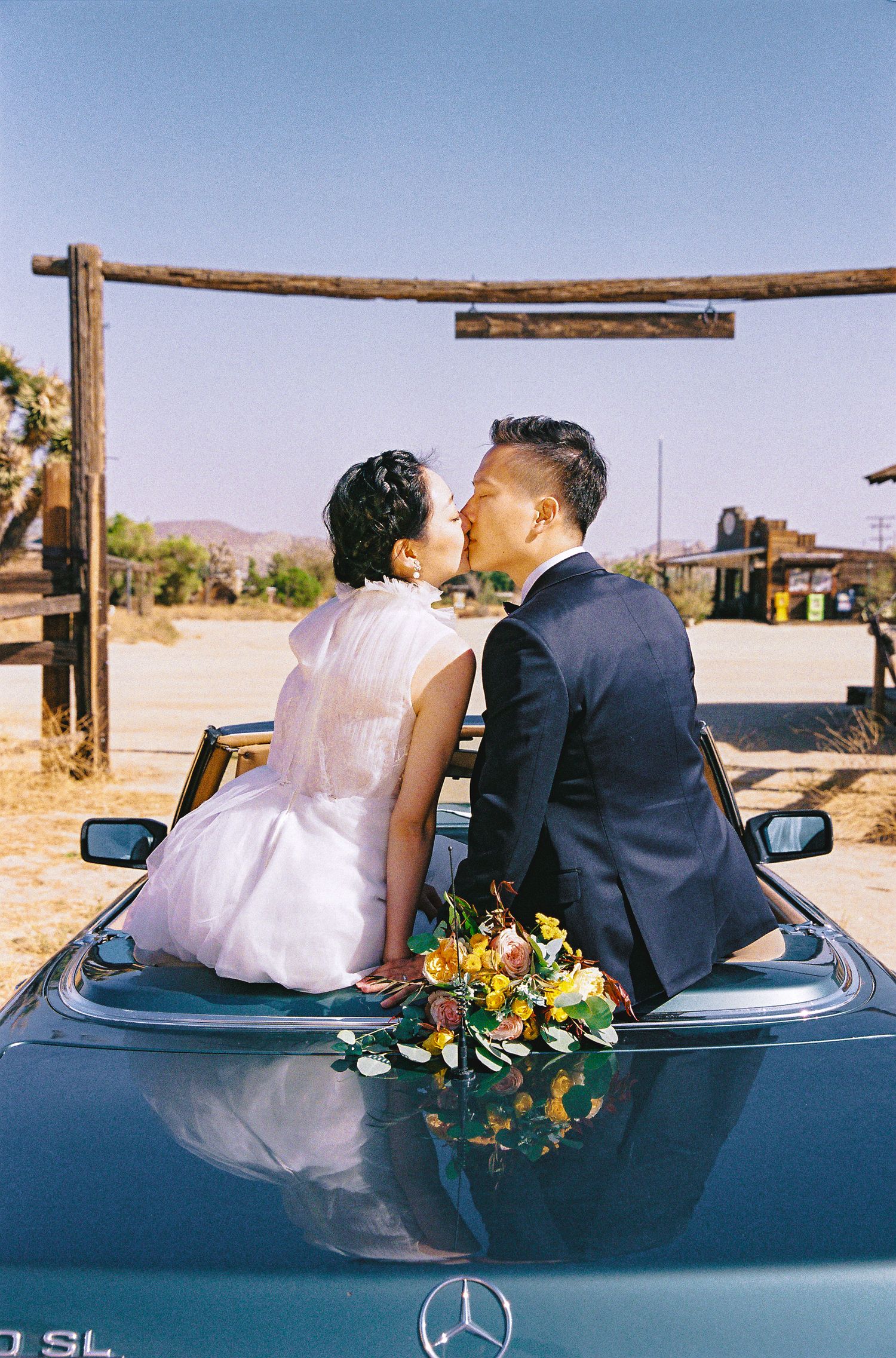 Newlyweds sharing a kiss while sitting on a Mercedes convertible in a desert setting.