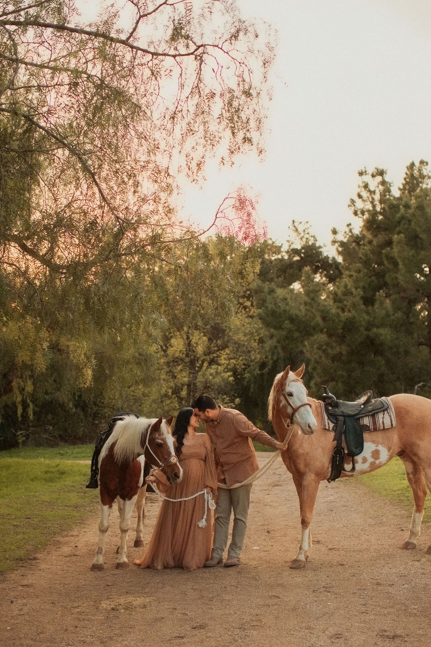 Two people with horses on a dirt path at sunset among trees casting warm golden light.