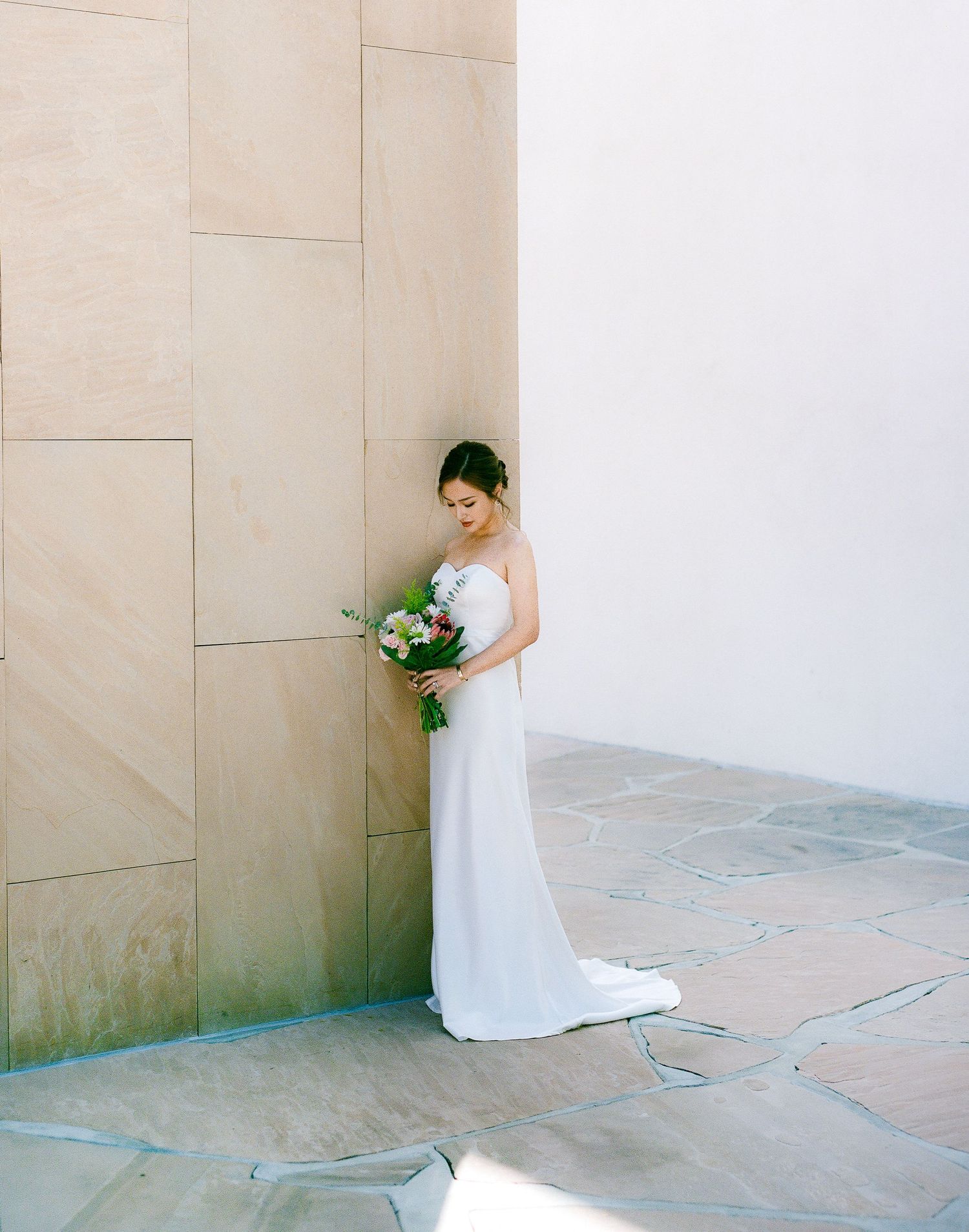 A bride in a white gown holds a bouquet while standing against a modern architectural wall.