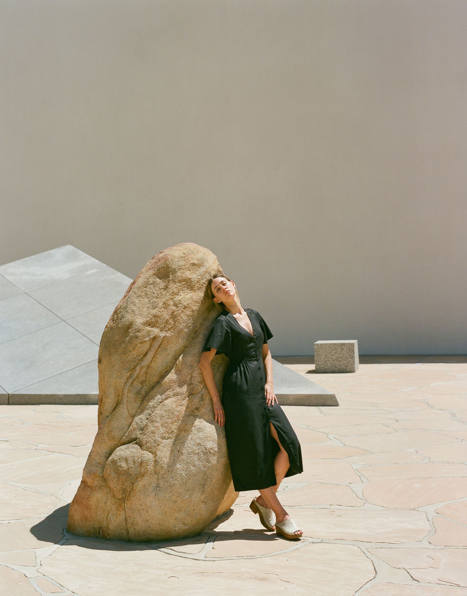 A fashion composition with a dark dress against a large sculptural rock in an outdoor setting.
