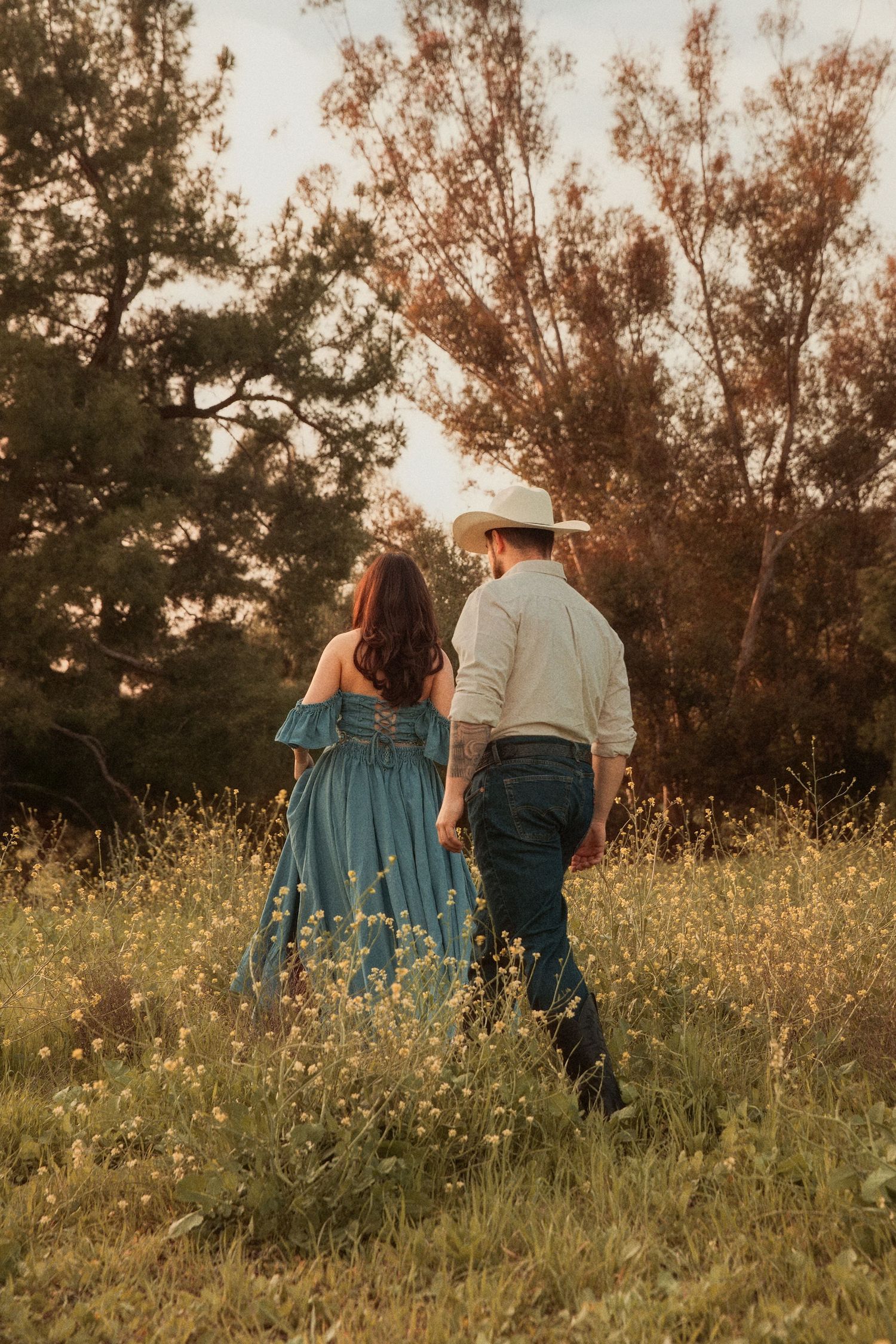 Couple in rustic attire walking through a wildflower meadow at sunset with tall trees in the background.