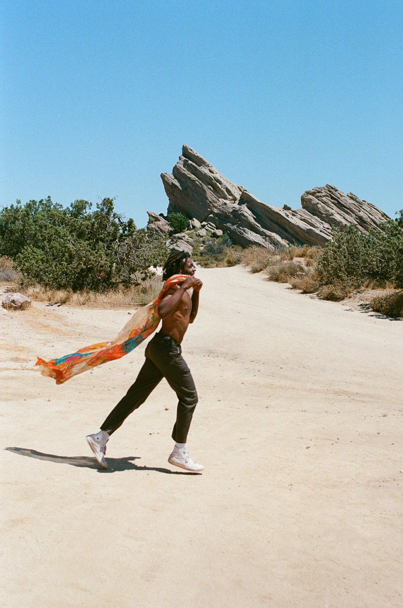 Person dancing with orange scarf on desert path with dramatic rock formations in background.