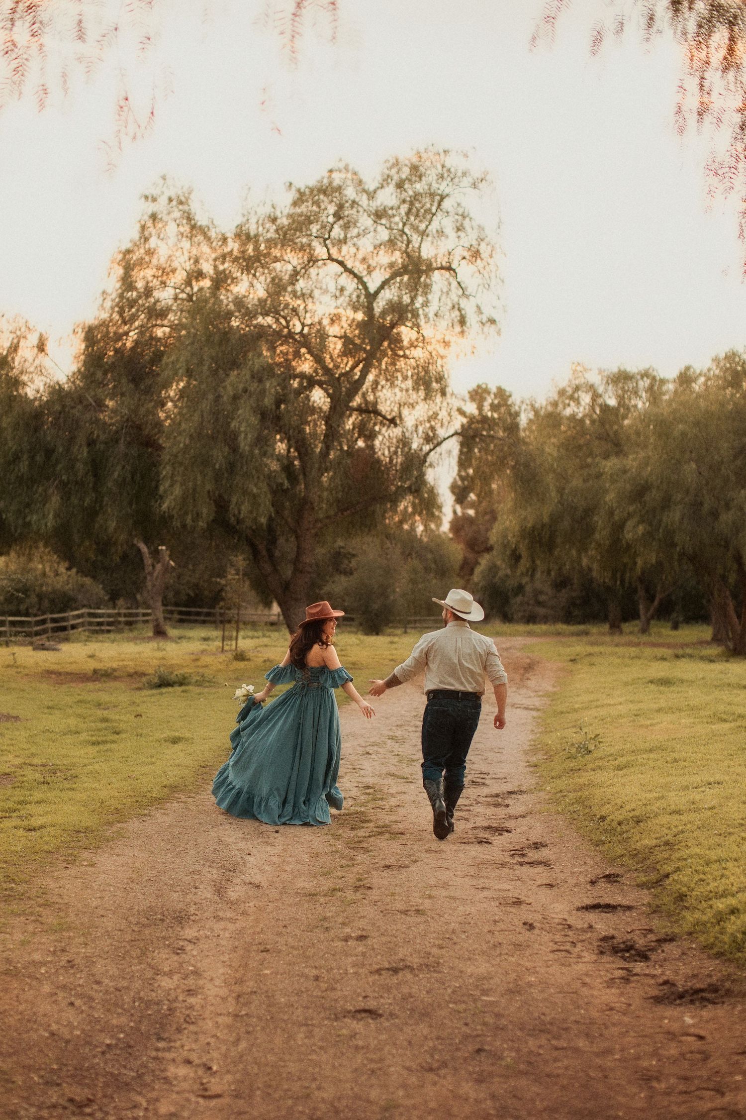 Two people in vintage clothing walk hand in hand down a dirt path through a dreamy sunset-lit field with willow trees.