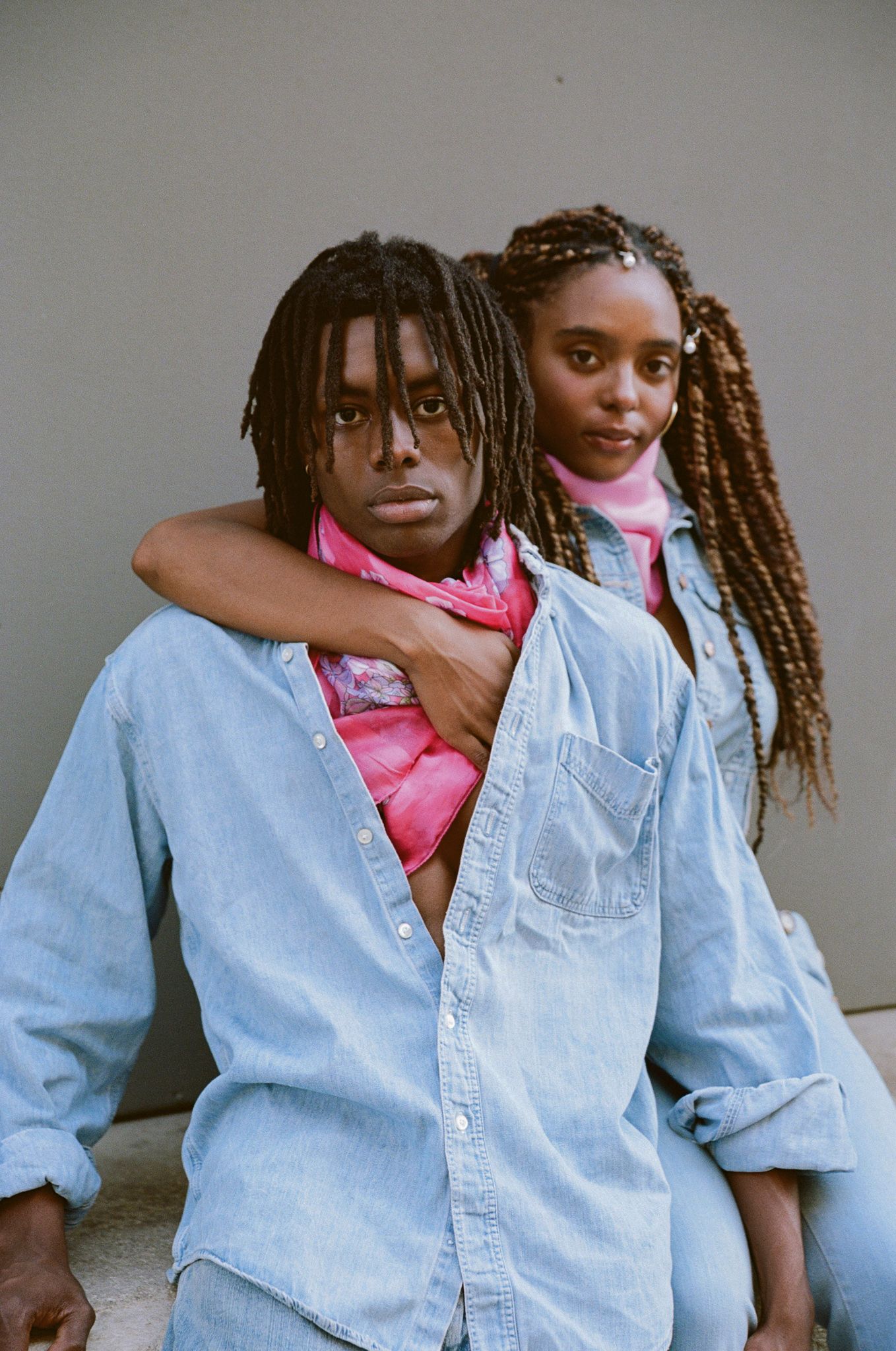 A pair wearing light denim shirts pose casually together against a neutral background.