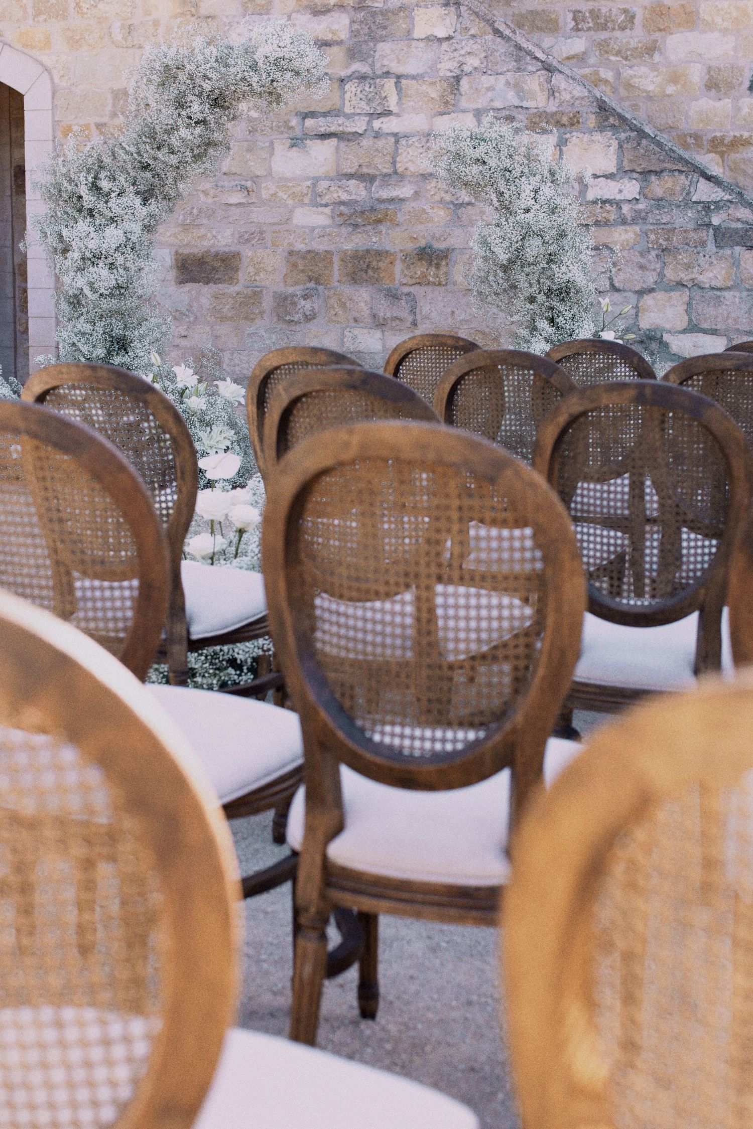 Rustic cane-back chairs arranged for a wedding ceremony with baby's breath floral arch against stone wall.