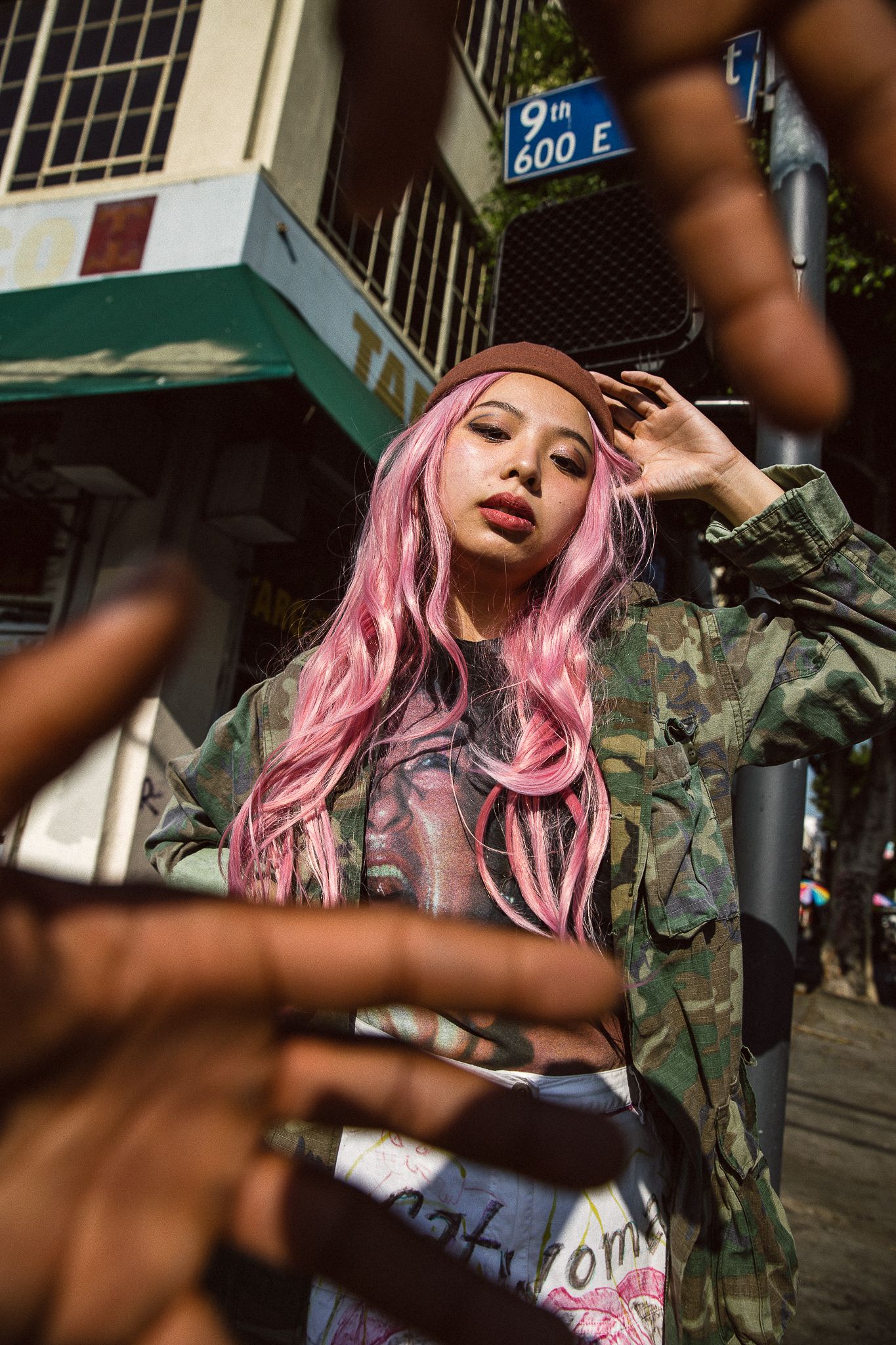 Model with pink hair in camo jacket poses on urban street corner.