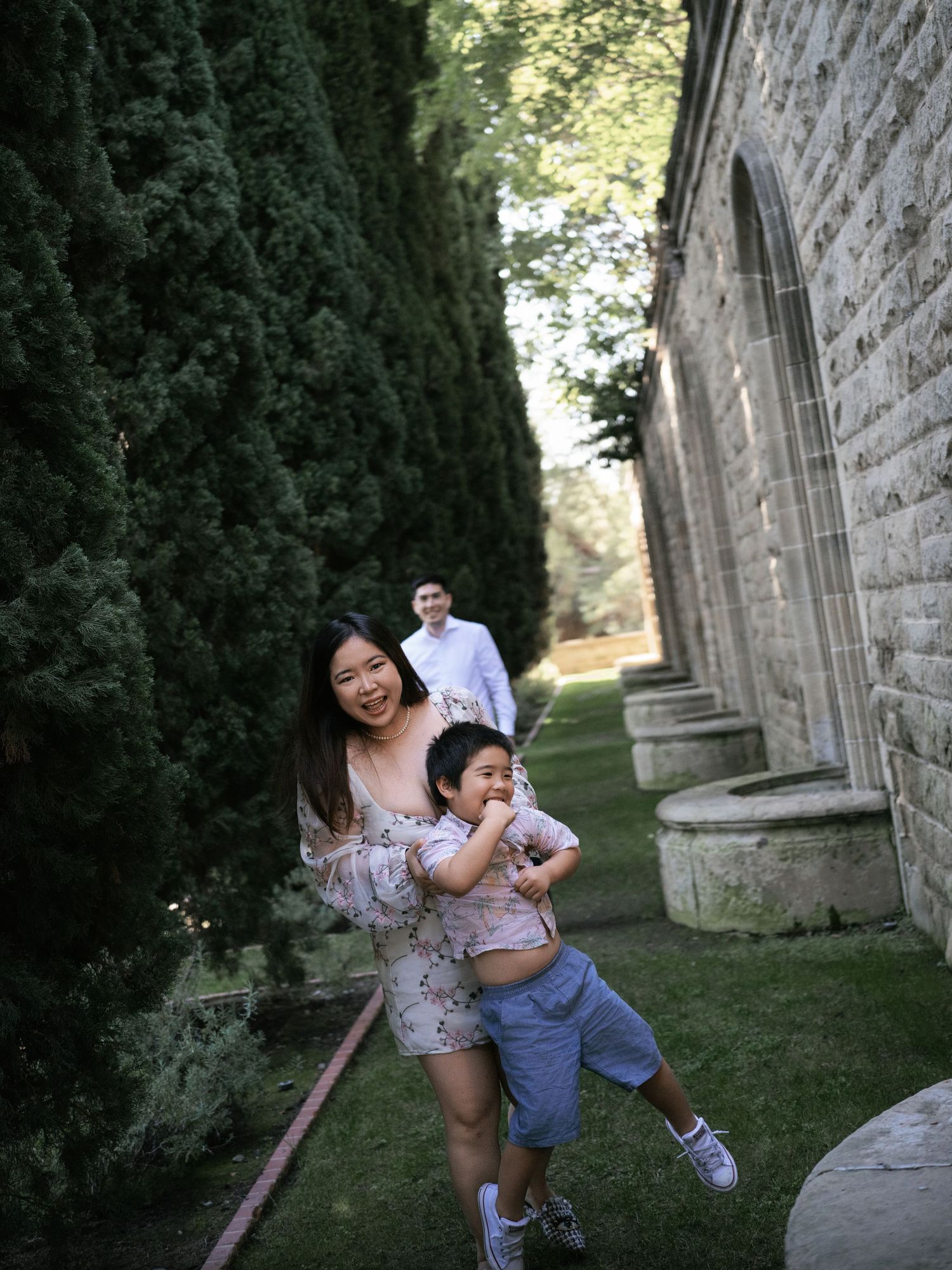 A playful family moment captured along a stone building with tall cypress trees.