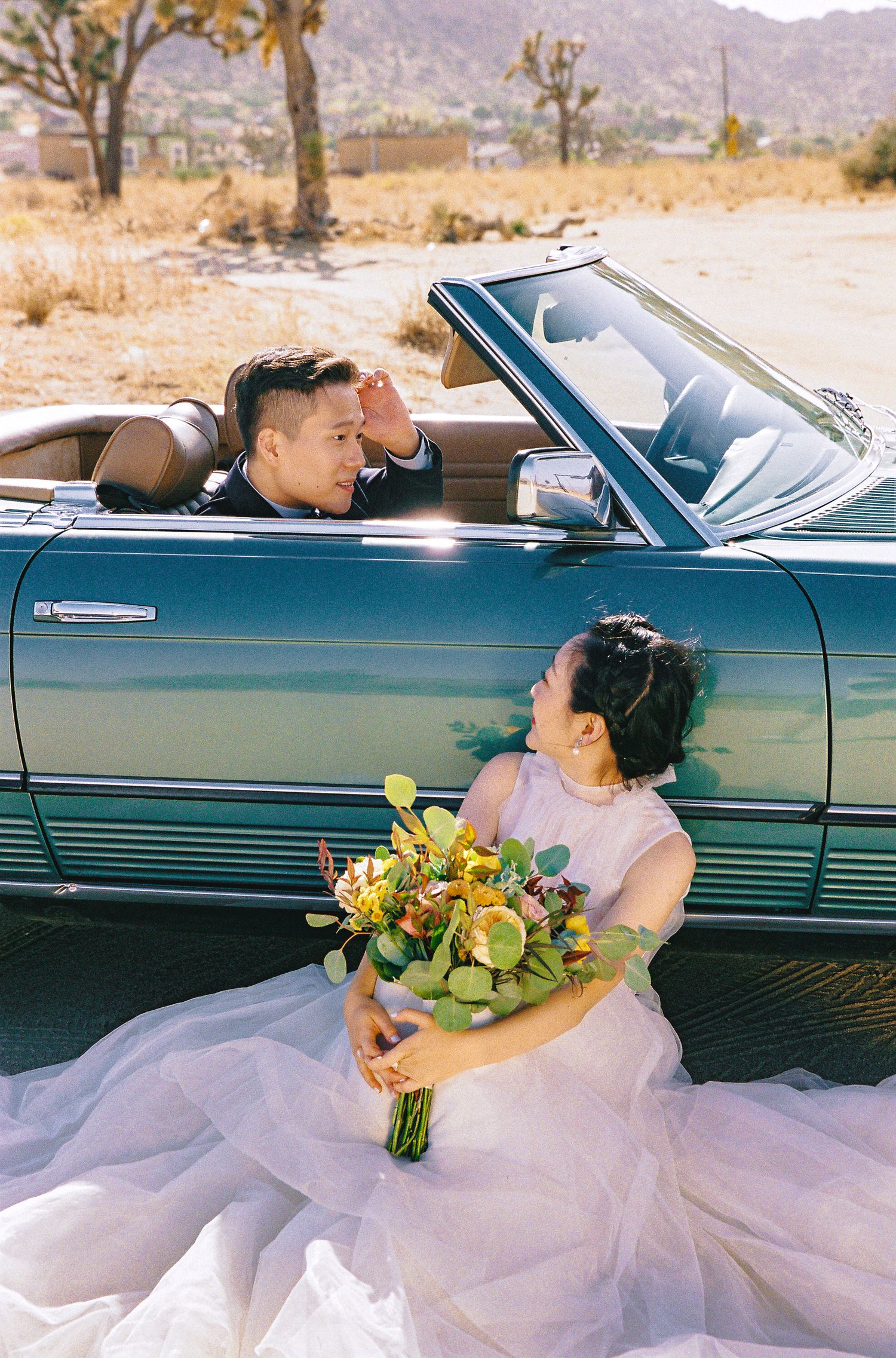 A couple poses with a vintage convertible in the desert, with a bouquet of yellow flowers and tulle dress.