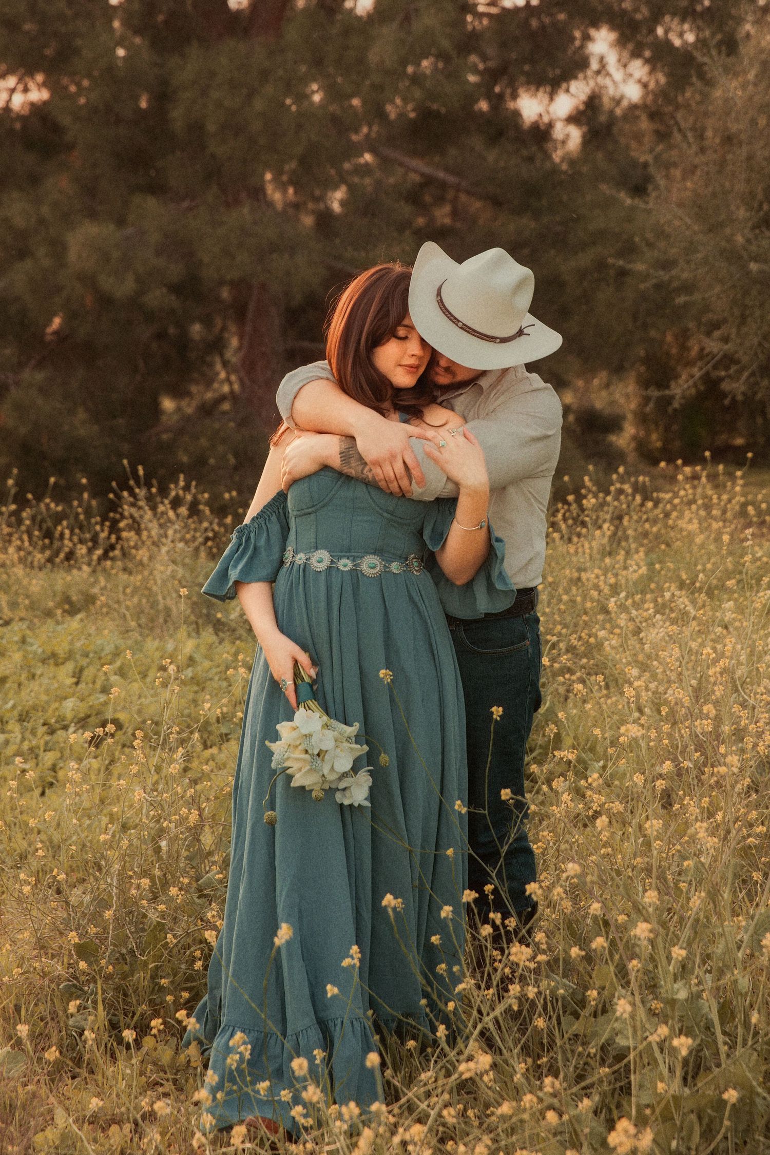 Romantic couple embraces in a wildflower meadow at sunset, woman in flowing teal dress holds a small bouquet.