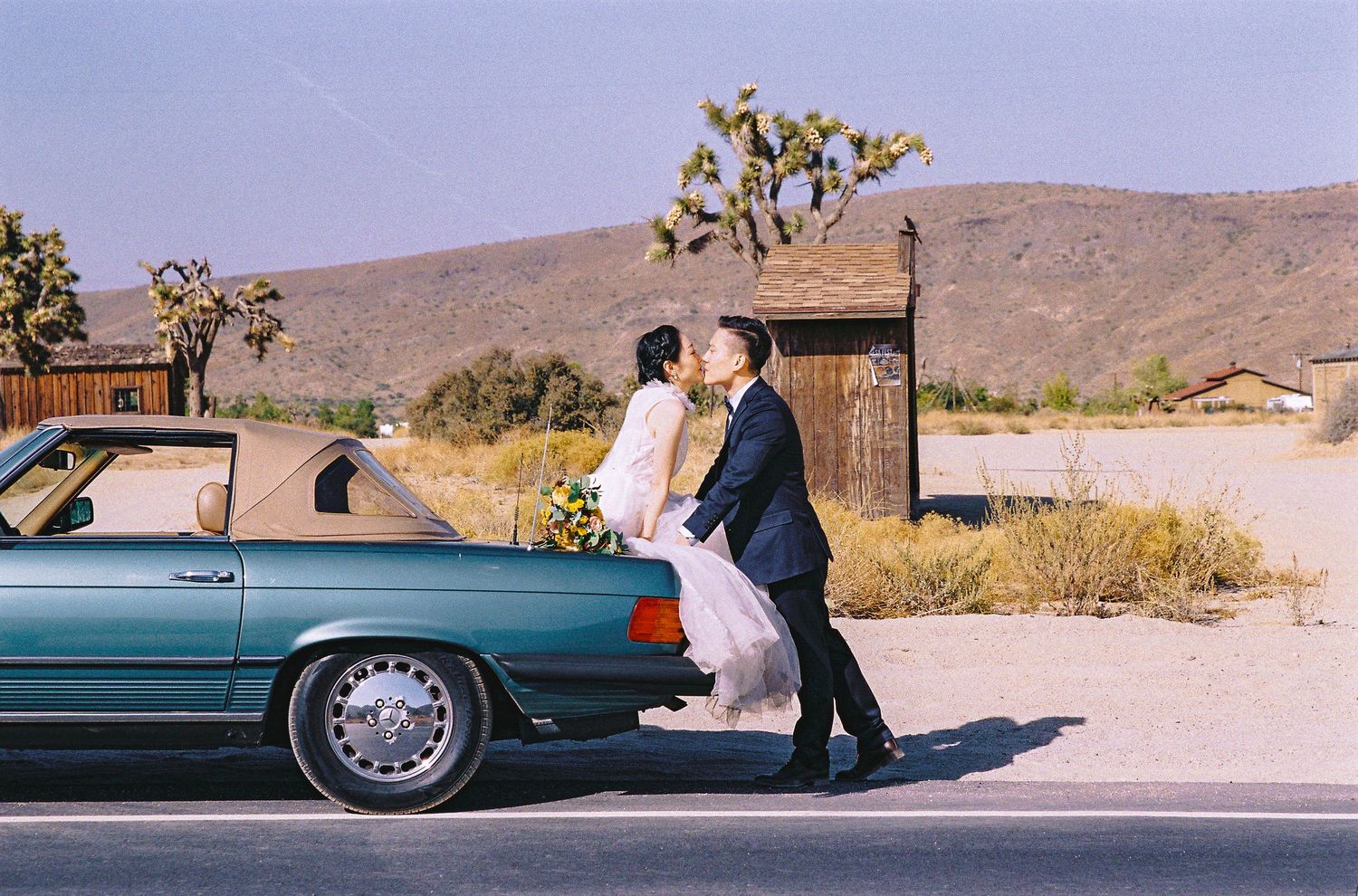 A couple shares an intimate moment against a teal vintage car in the desert landscape.