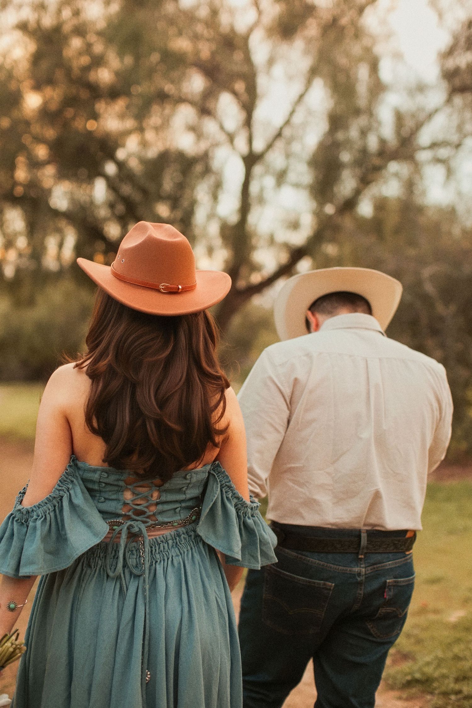 A couple in western attire walk together, wearing cowboy hats with the woman in a blue dress at golden hour.