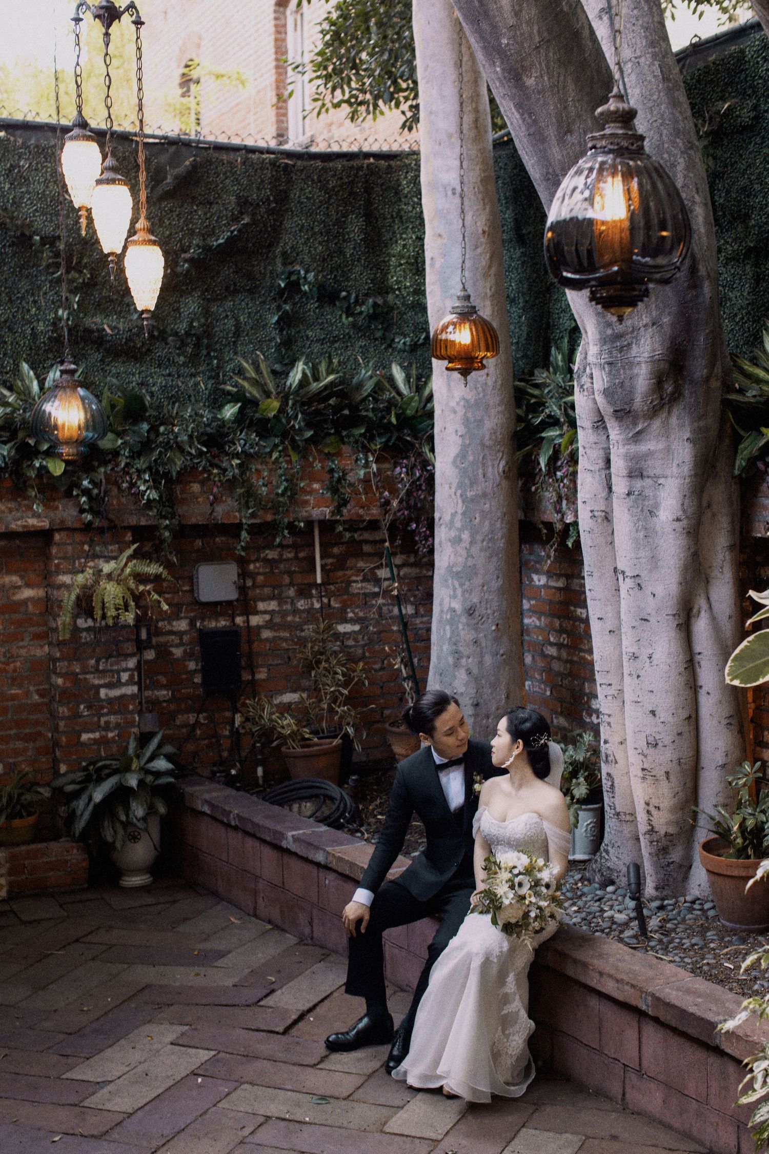 Couple sits on brick bench under hanging lanterns in garden courtyard.
