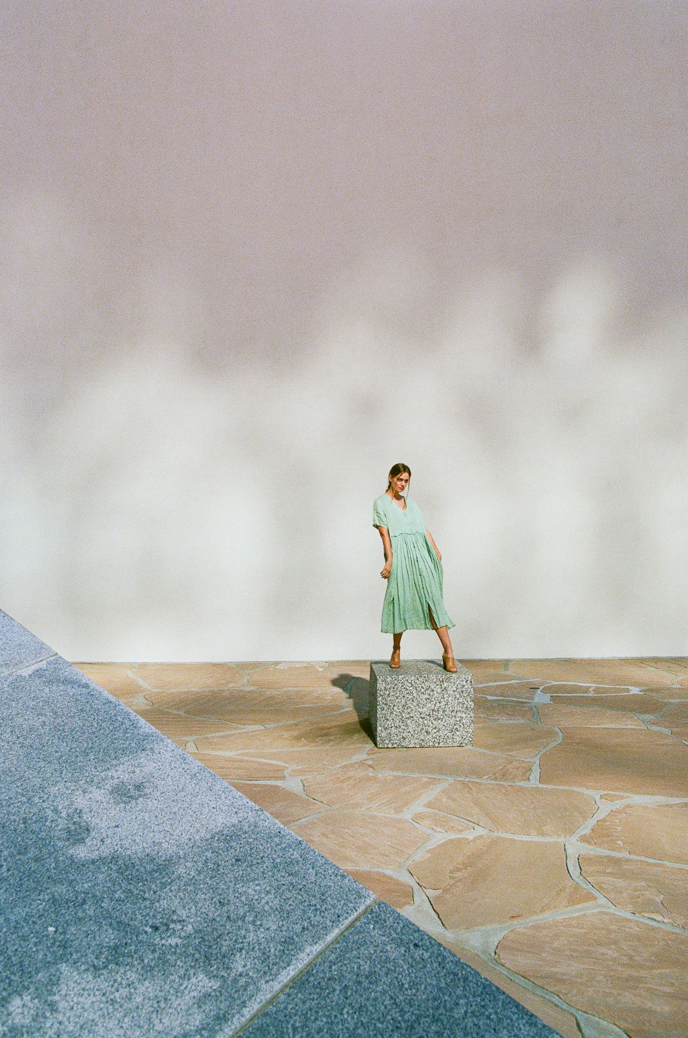 Artistic shot of figure in mint green dress standing on concrete block against geometric backdrop.