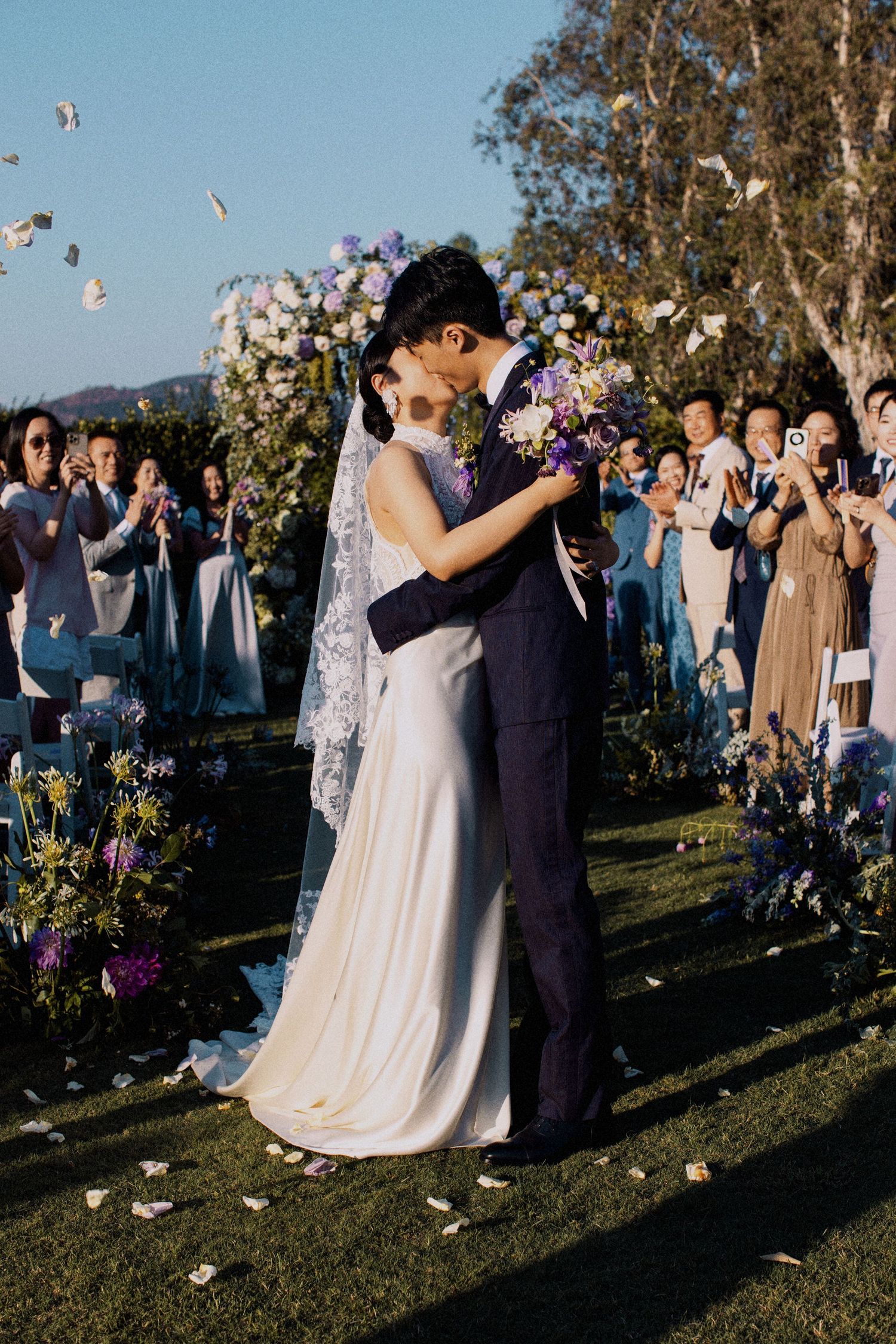 Newlyweds share their first kiss as guests throw flower petals in an outdoor garden setting.