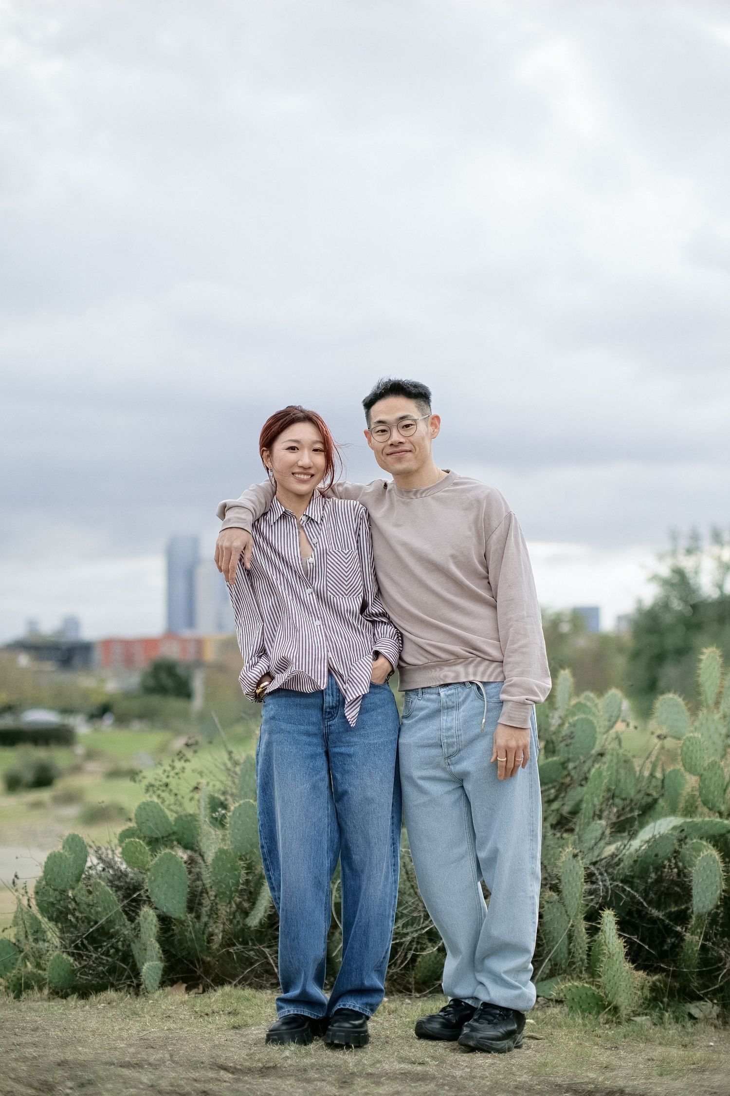 Two people stand together among cactus plants with a cloudy city skyline in the background.