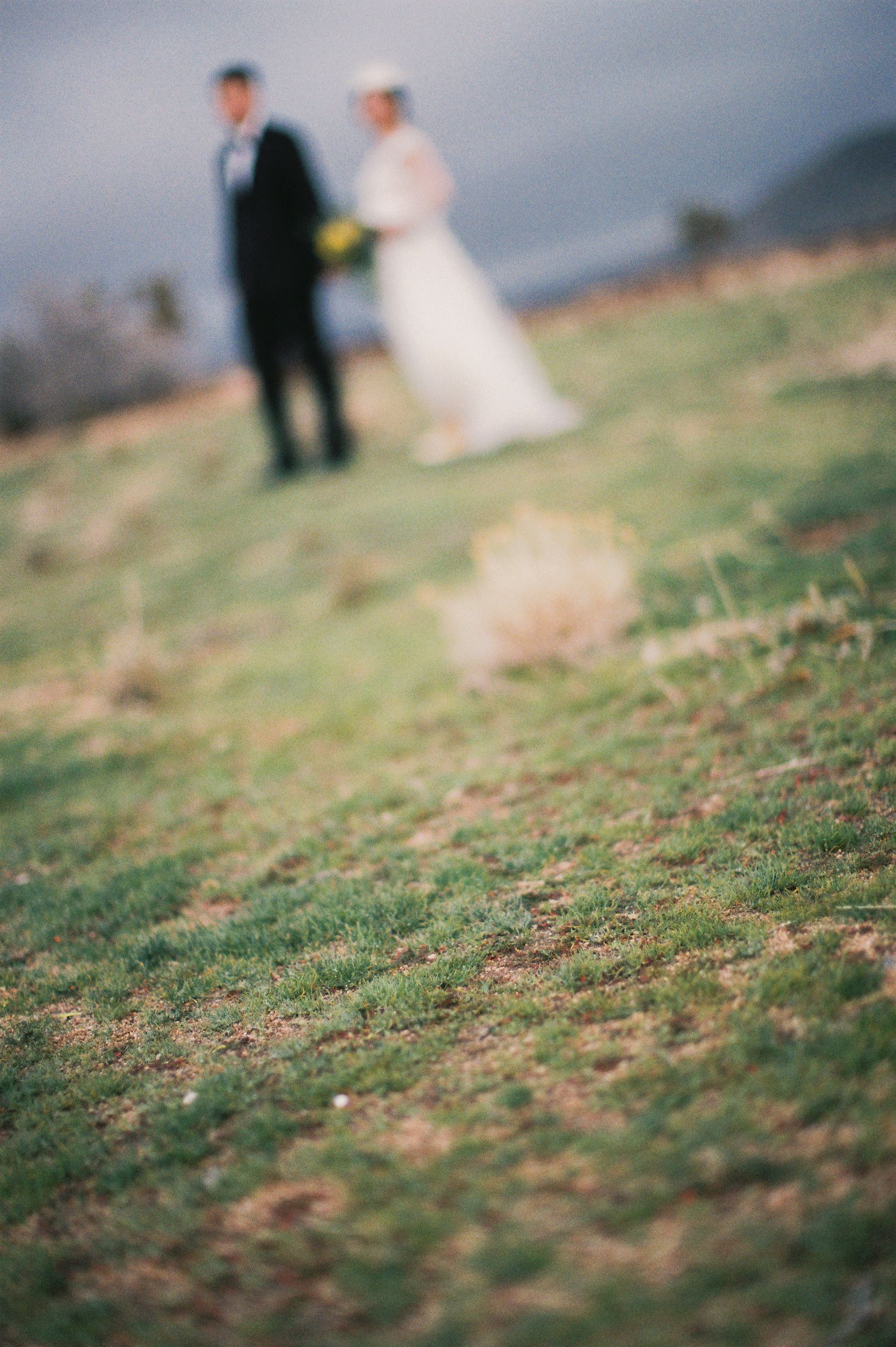 Dreamy out-of-focus wedding photo with couple standing on hillside against cloudy sky.