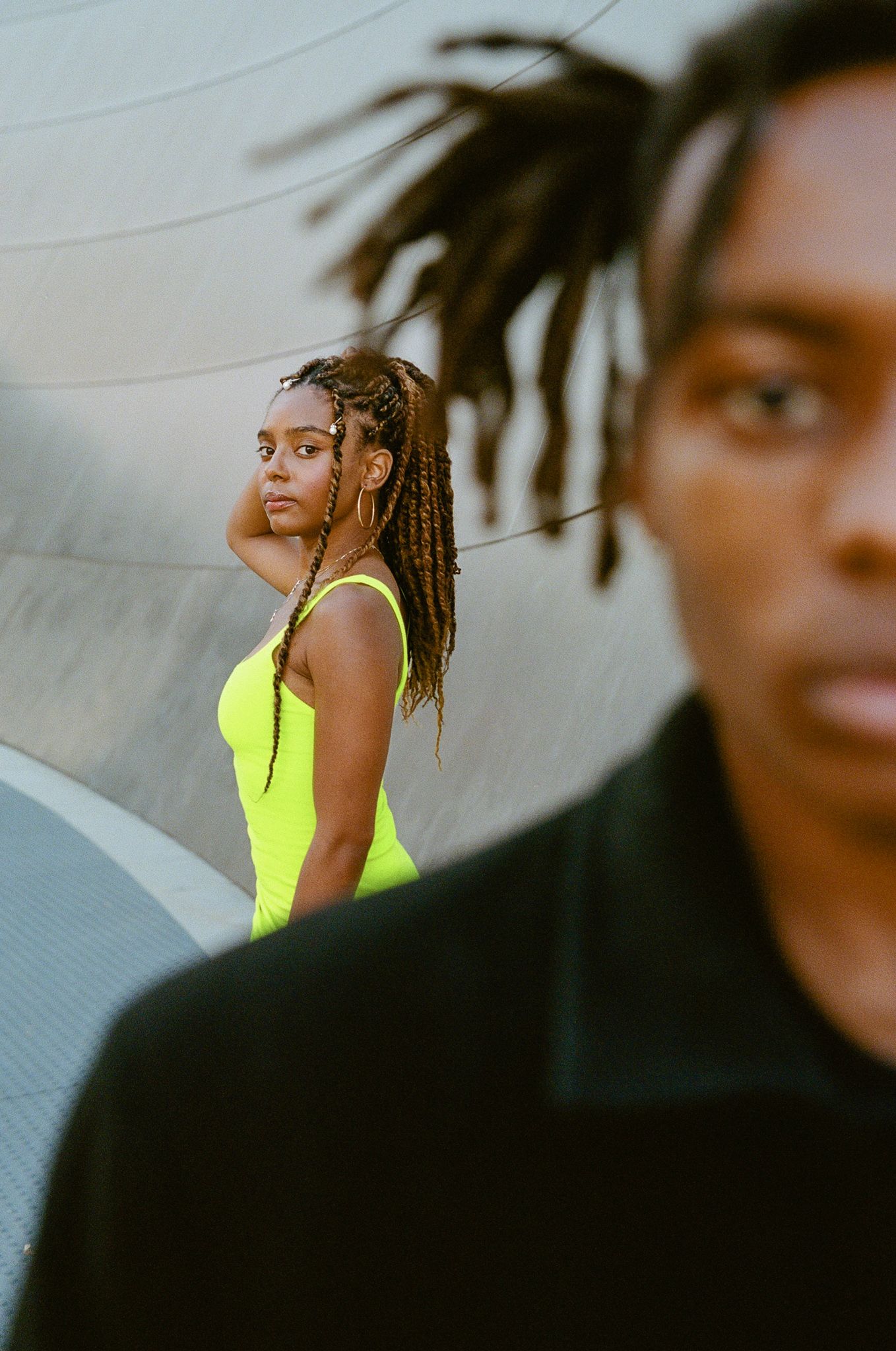 Person in neon yellow top stands in contrast against dark background while another figure is blurred in foreground.