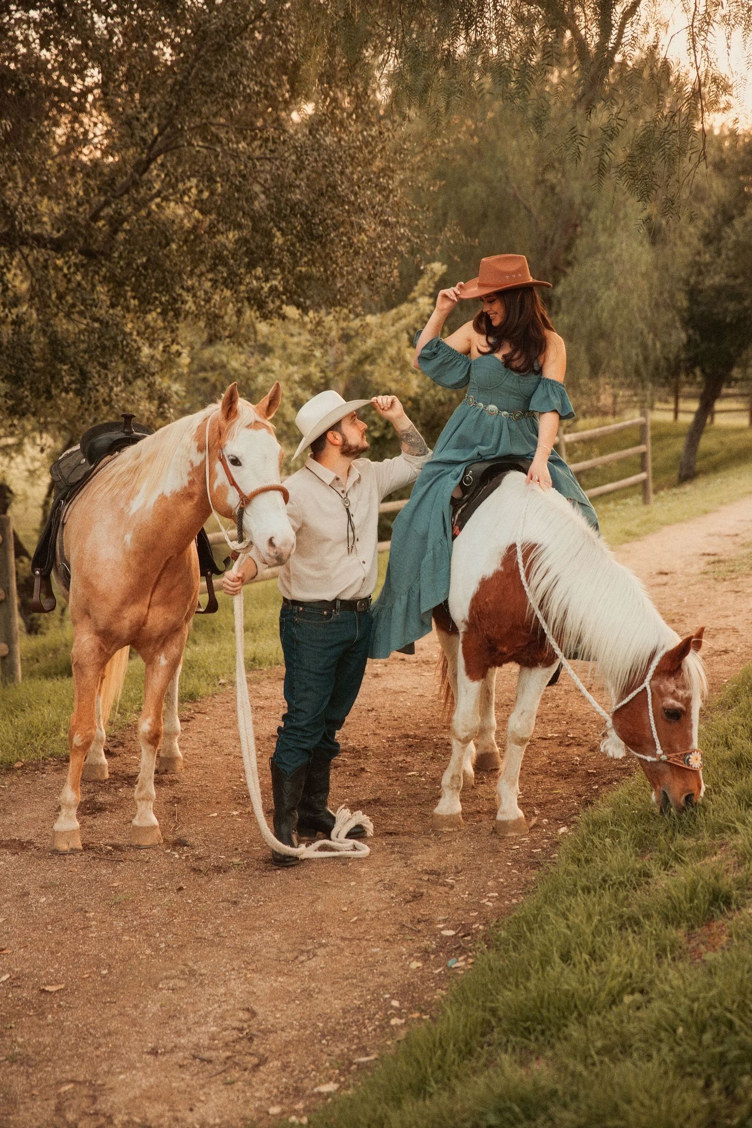 A couple with horses at sunset on a rural path with wooden fencing and trees in the background.