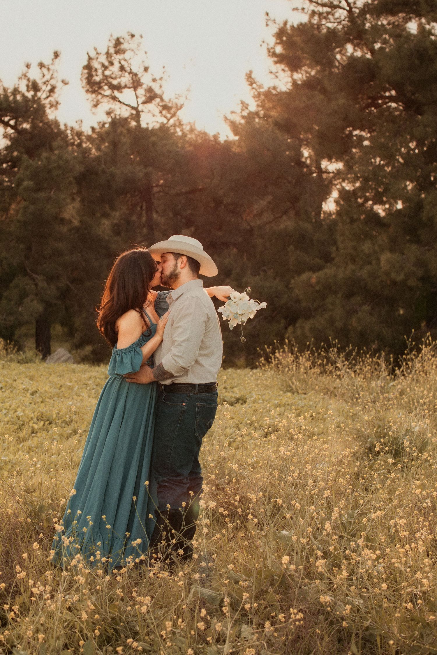 A couple embraces in a golden field at sunset with pine trees in the background.