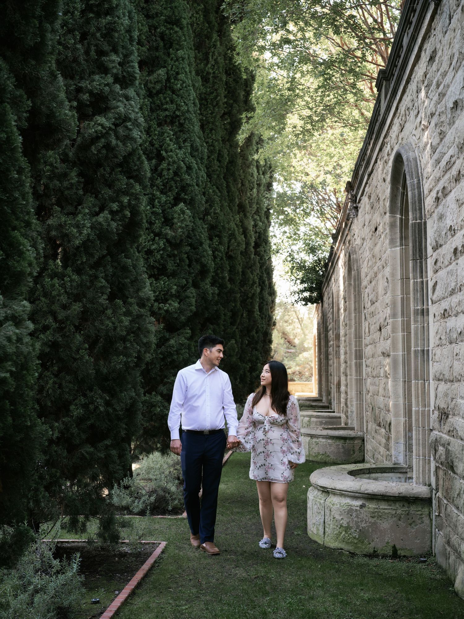 A couple strolls along a stone church building with cypress trees creating a romantic garden walkway.