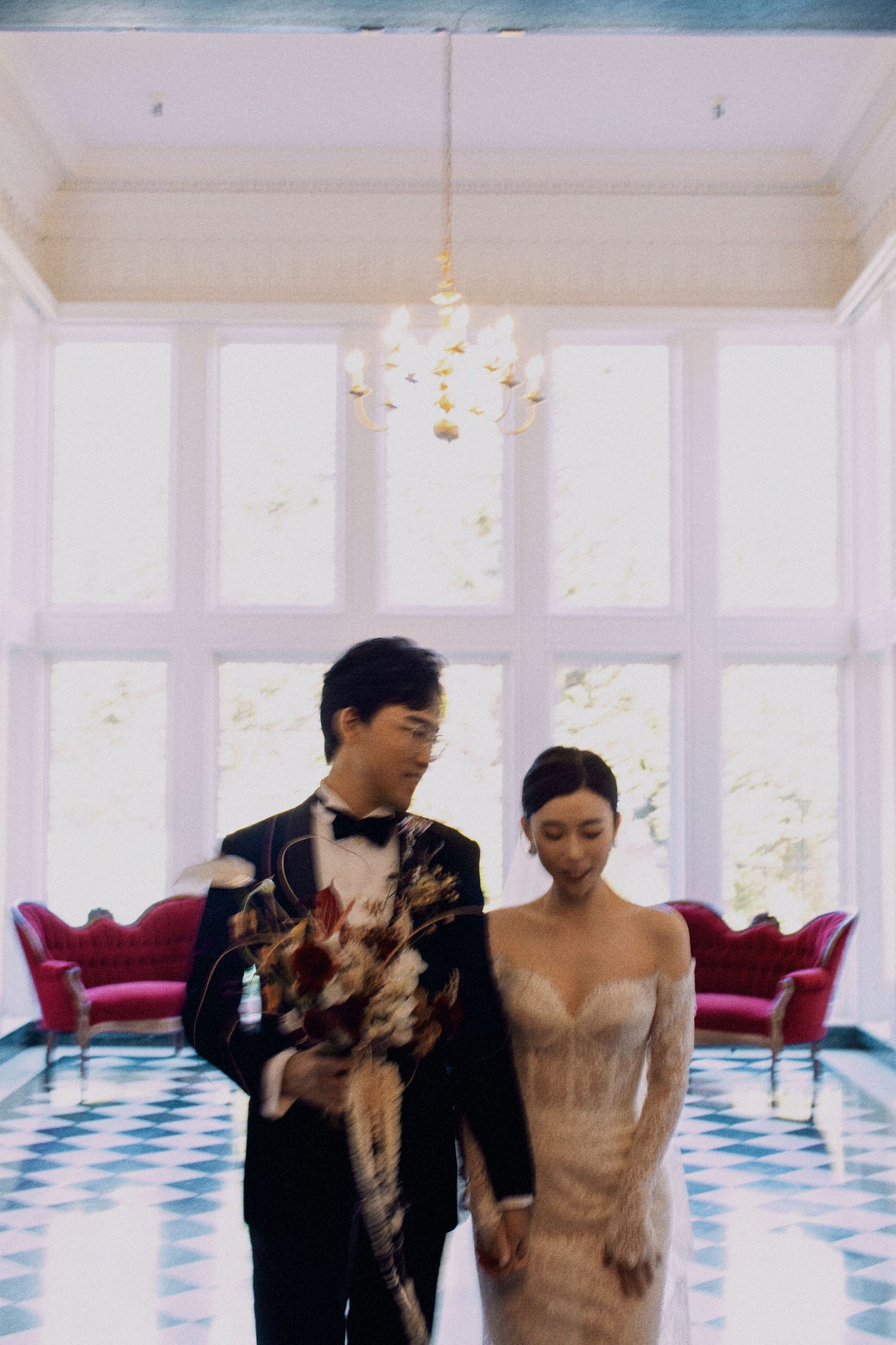A couple poses with their small dog in an elegant room with a chandelier and red velvet seating.