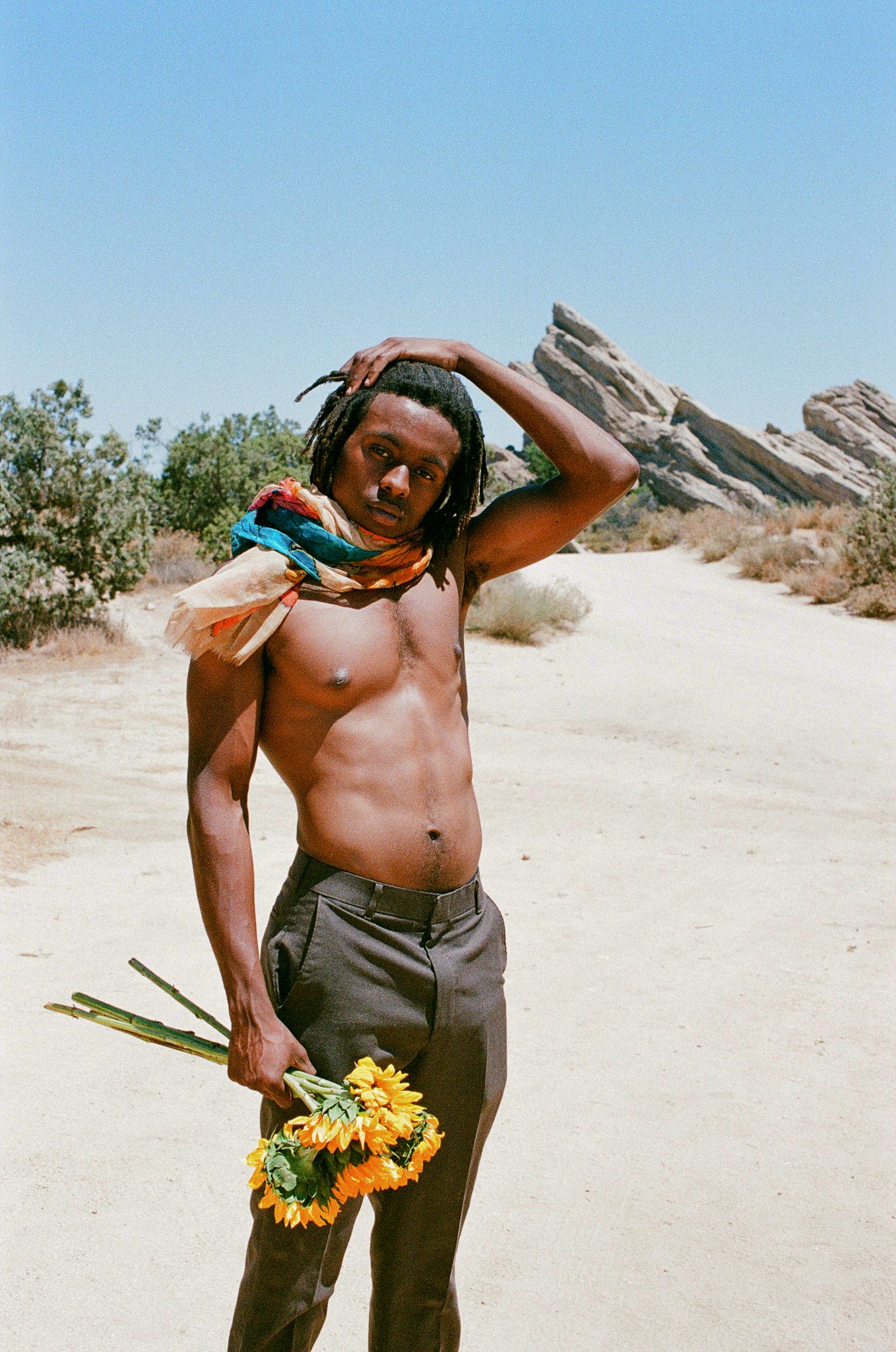 Person holding sunflowers poses on desert trail with rocky outcrop in background.