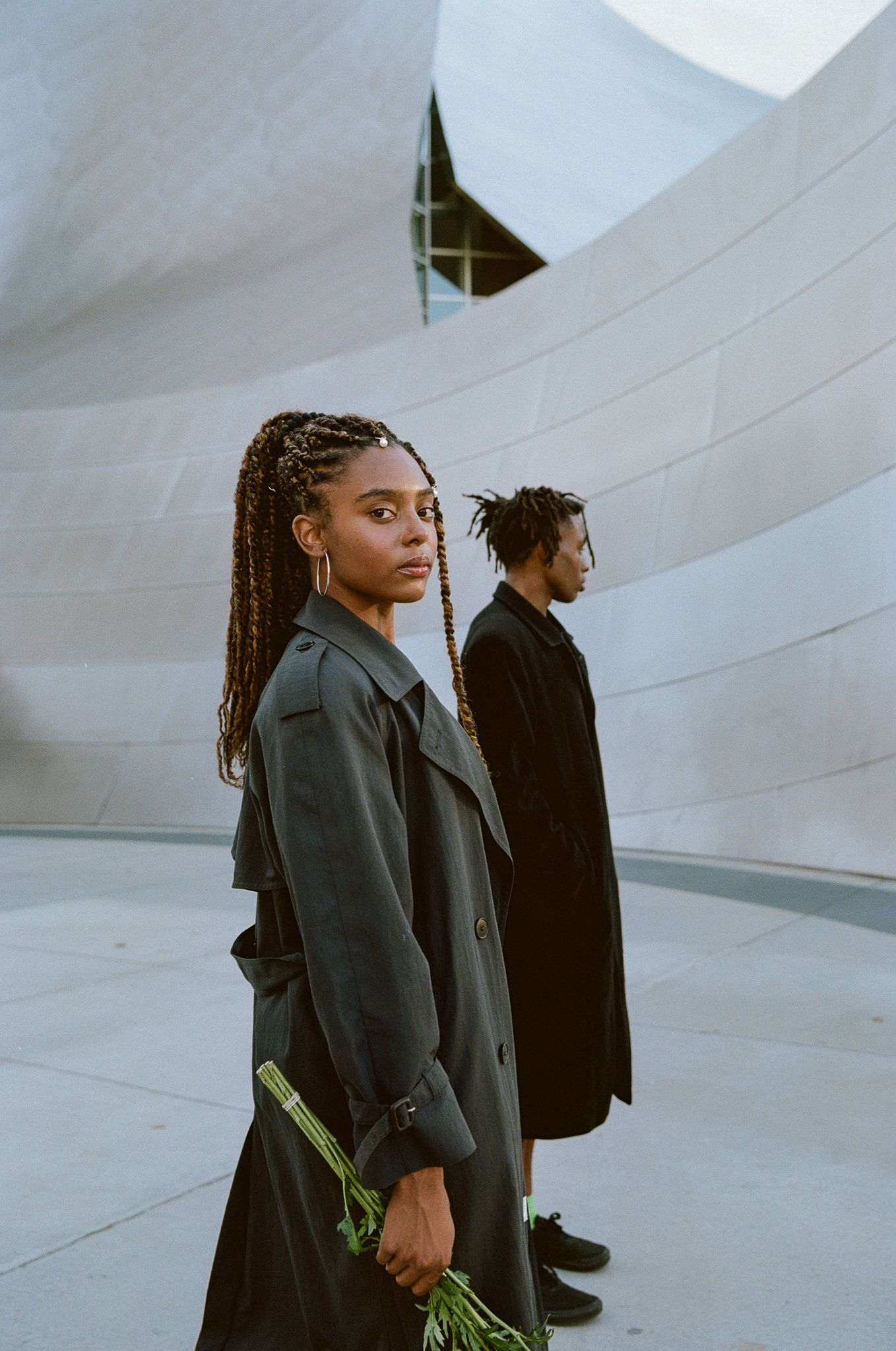 Two people in dark coats pose against curved white architectural wall during artistic fashion photoshoot.